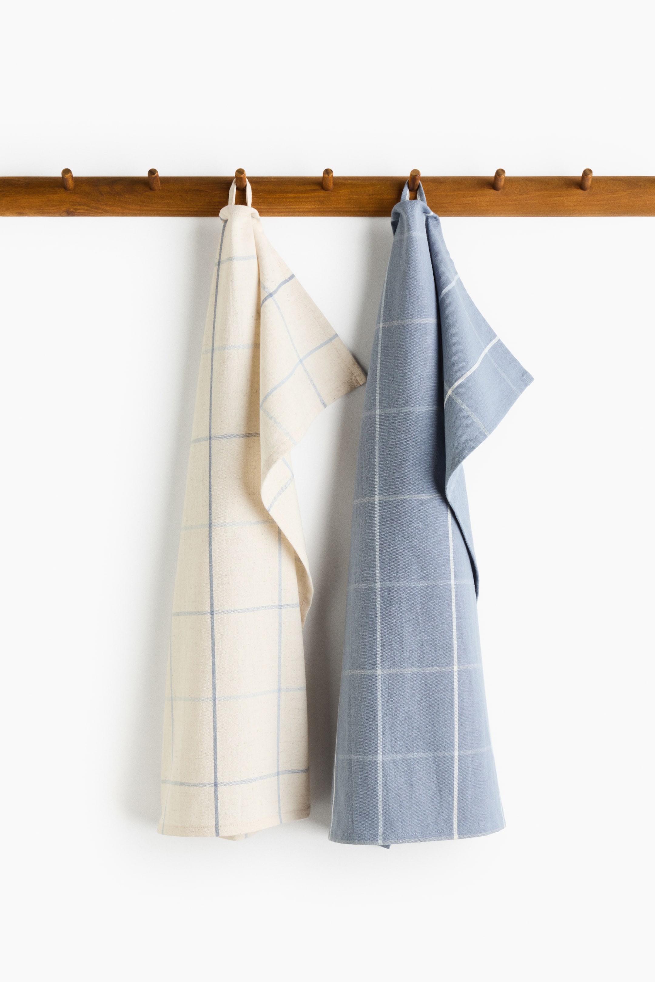 View larger image: Tea towels are hanging, with one cream towel displaying a blue yarn-dyed grid pattern and one light blue towel displaying a white yarn-dyed grid pattern. Both feature a cotton and linen weave and a hanger loop on one short side.