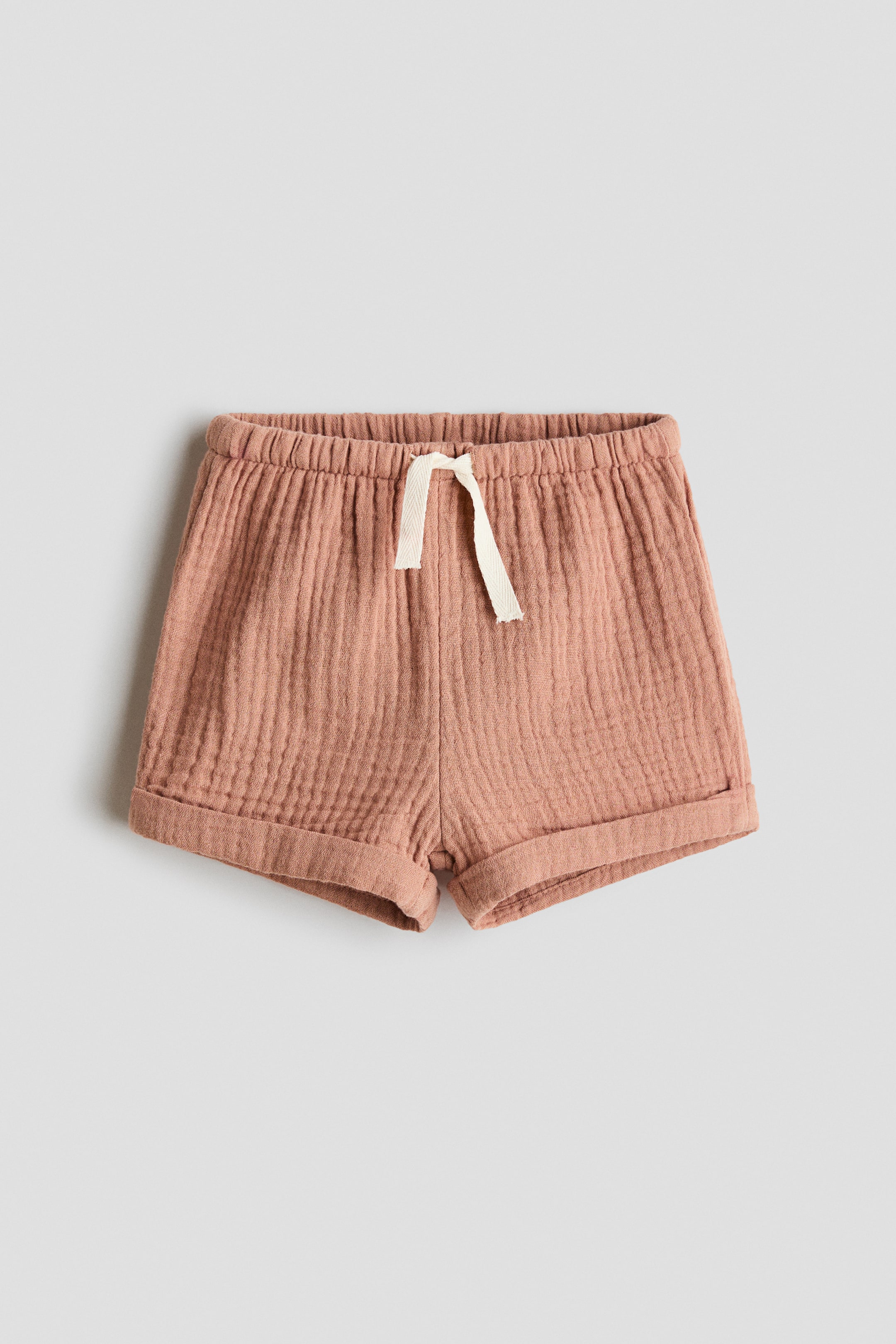 View larger image: Muted pink muslin shorts come with an elasticated drawstring waist, showcasing a light-coloured tie at the front. The hems feature sewn-in turn-ups for added style.