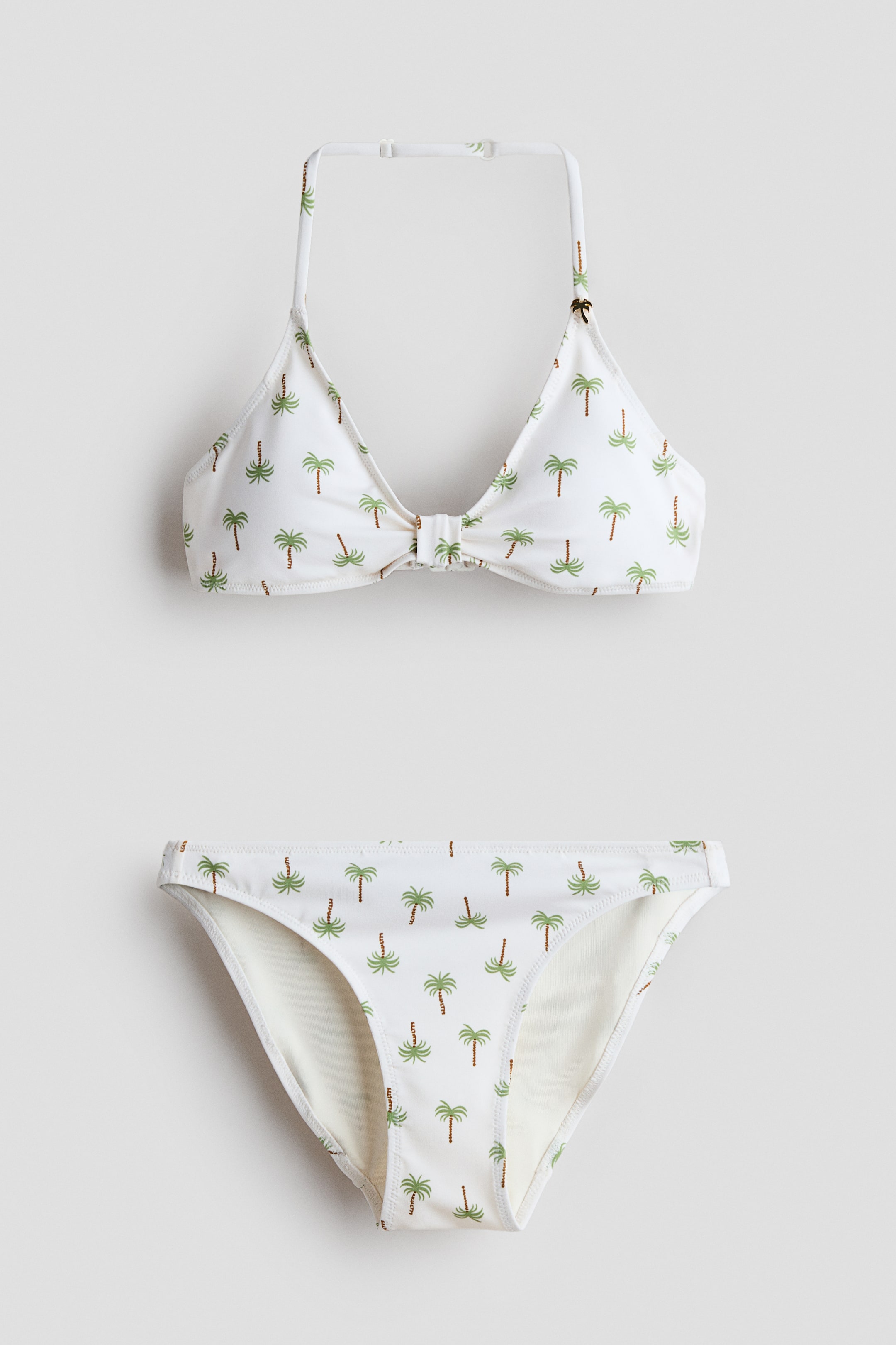 Ver imagen más grande: Un conjunto de bikini de dos piezas presenta una tela blanca con un patrón repetido de palmeras verdes y marrones. La parte superior de cuello halter muestra copas triangulares con un detalle de nudo central, y la parte inferior a juego presenta un corte clásico de braguita.