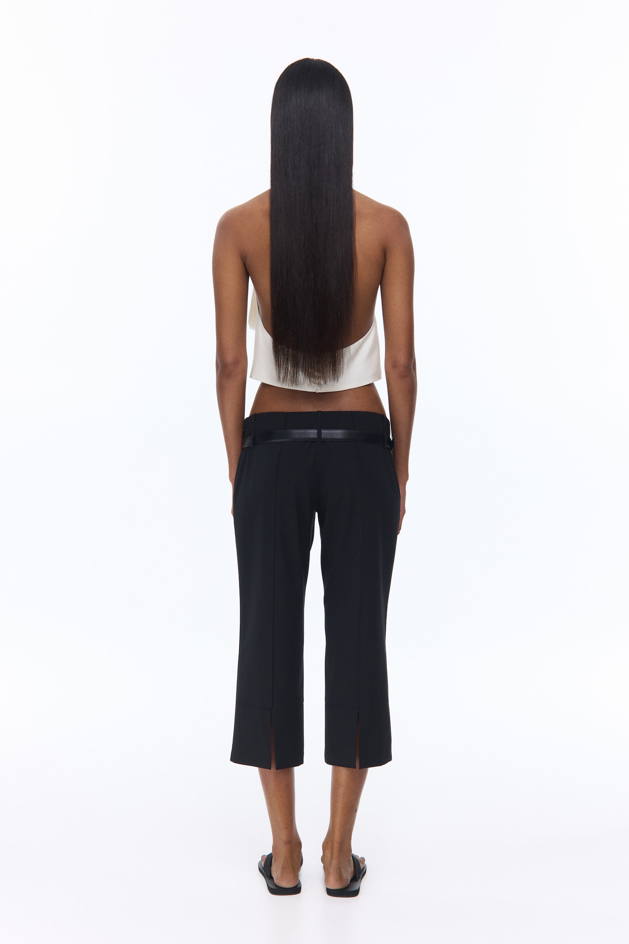 View larger image: The model is showcasing black 3/4-length trousers with a low waist and straight legs. A black belt is elegantly threaded through the double belt loops. The design features sewn-in creases running down the back, complemented by small slits at the back of the hems.