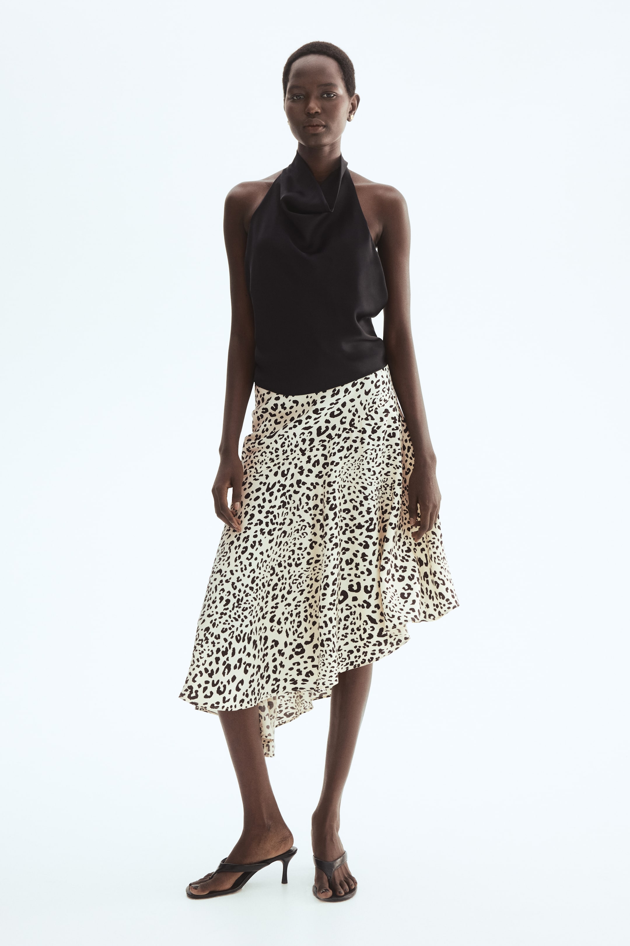 View larger image: A black halter neck top featuring a cowl neck is layered with a cream and black leopard print skirt. The skirt showcases an asymmetrical hem and a flowing, draped design.