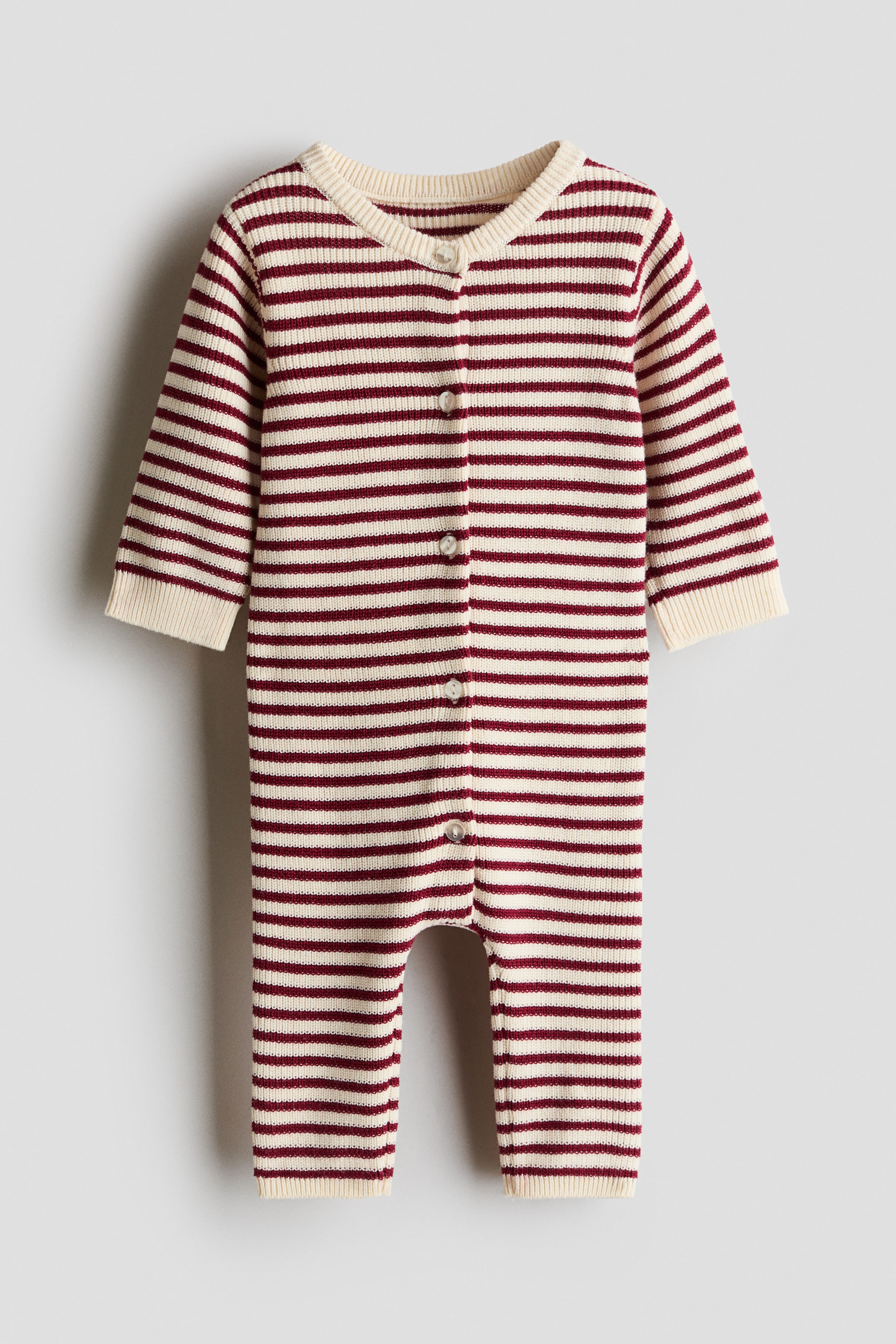 View larger image: A striped baby knit jumpsuit is shown from the front, displaying a pattern of horizontal red and cream stripes. The long-sleeved garment features a central button fastening with small white buttons and is finished with cream-coloured ribbed cuffs at the neckline, wrists, and ankles.