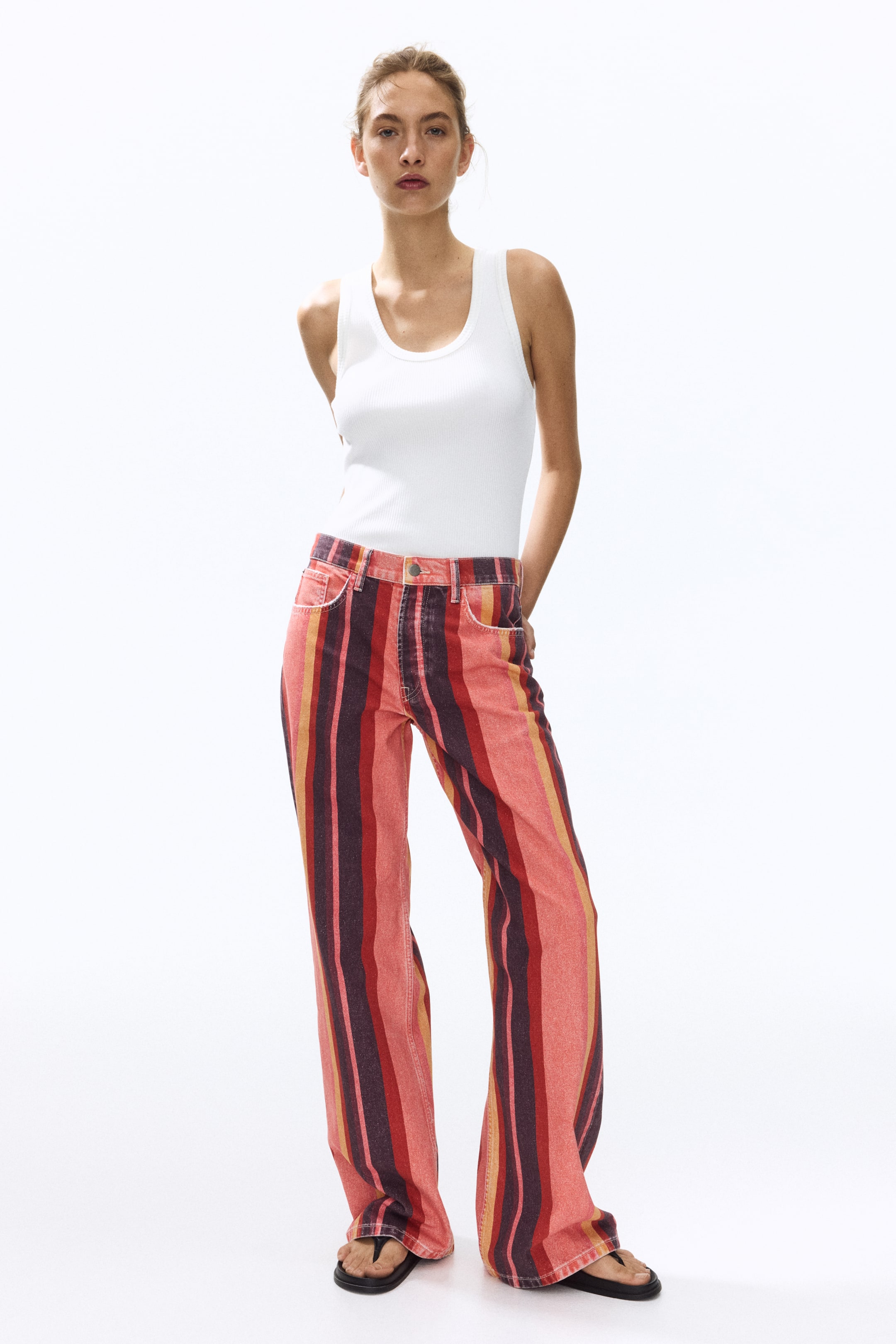 Ampliar la imagen: La modelo lleva puestos unos pantalones de mezclilla de algodón con 5 bolsillos. Los pantalones presentan una cintura regular con cierre de cremallera y botón, piernas rectas, y un patrón de rayas verticales en tonos de rojo, naranja y marrón. La modelo está colocando sus manos en los bolsillos frontales de los pantalones.