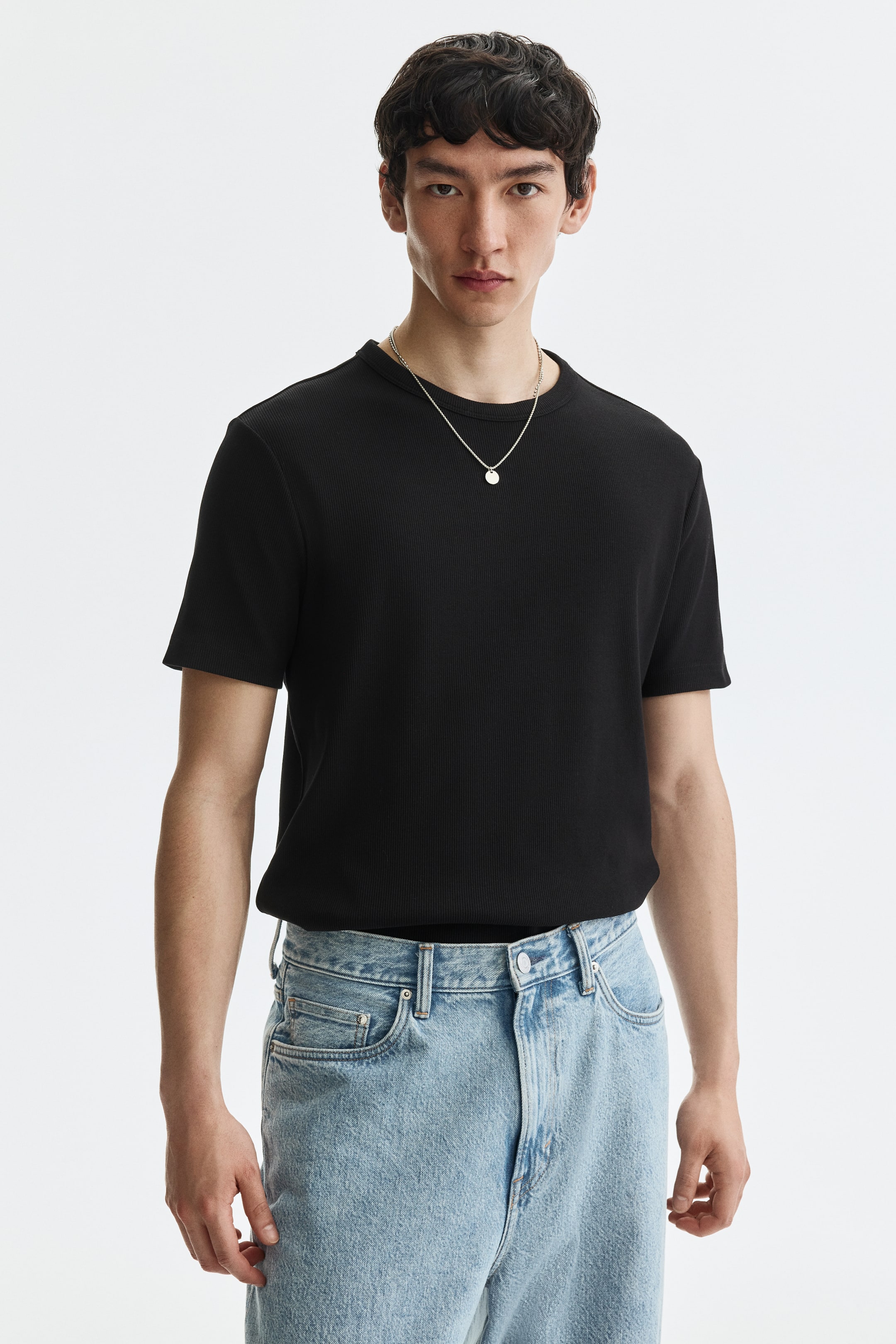 View larger image: The model is wearing a black ribbed t-shirt crafted from cotton jersey. It showcases a round neckline, short sleeves, and a straight cut hem, offering a sleek fit. A silver-toned necklace with a circular pendant elegantly rests on the t-shirt.