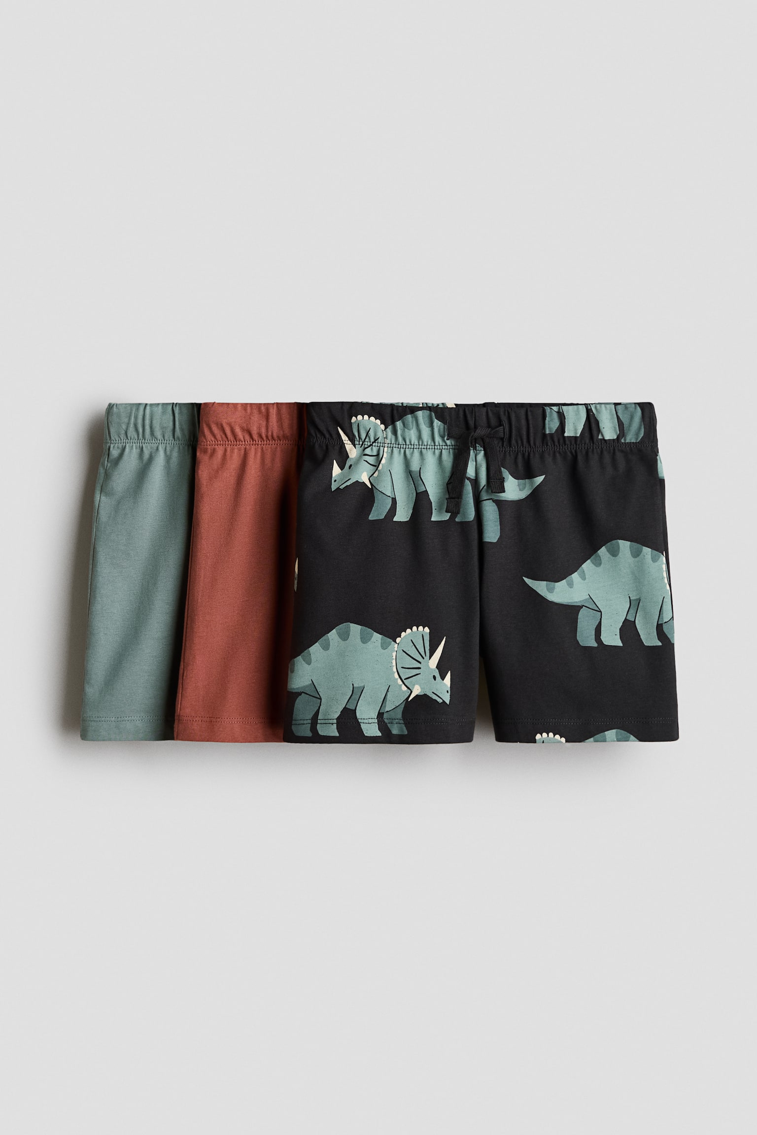 A three-pack of cotton jersey shorts featuring a solid sage green pair, a solid rust-red pair, and a black pair with a light green triceratops pattern. The shorts are shown from the front, with elasticated waistbands, and the front pair includes a small drawstring tie at the centre.