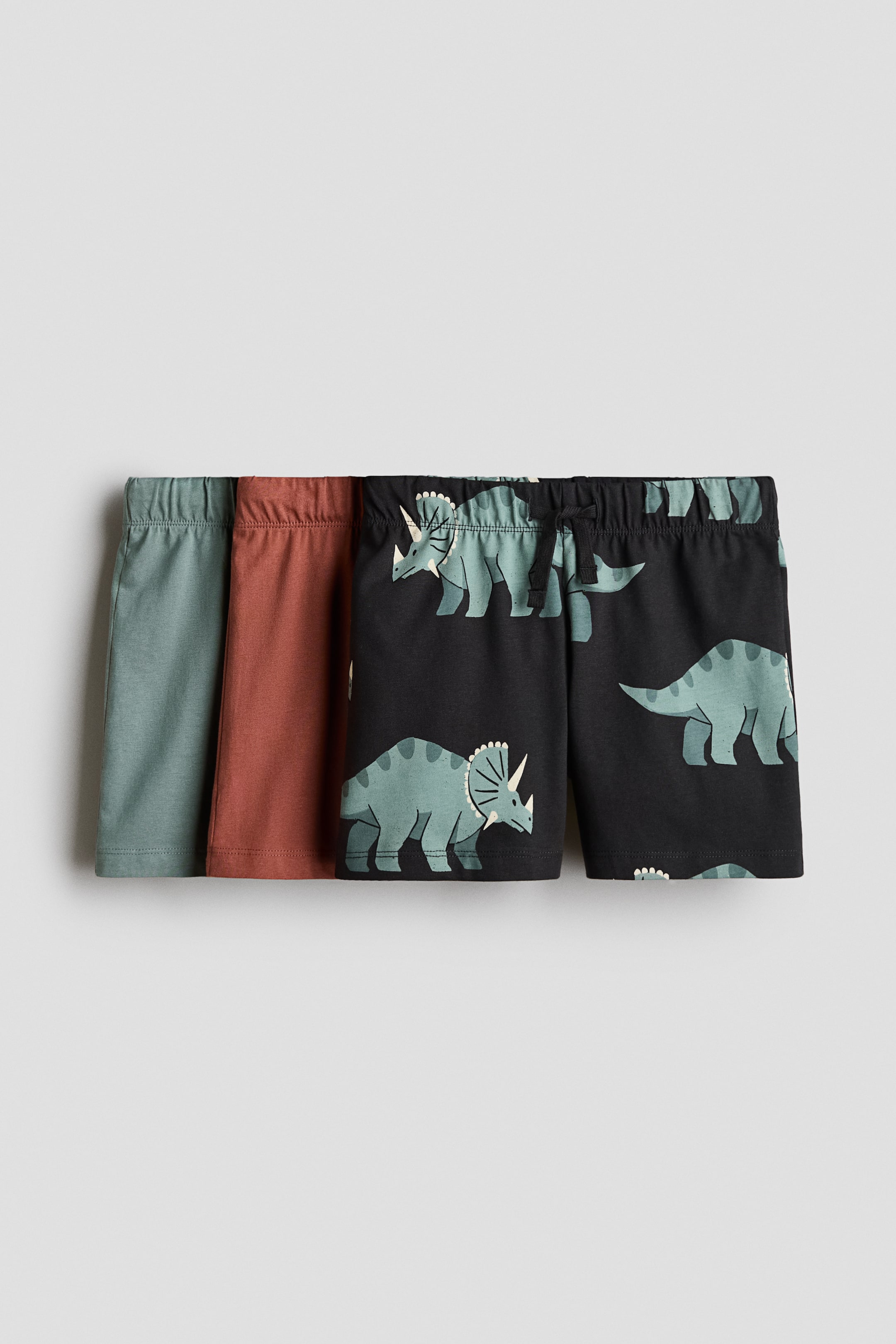 View larger image: A three-pack of cotton jersey shorts is displayed, featuring a solid sage green pair, a solid rust-red pair, and a black pair with a light green triceratops pattern. The shorts are shown from the front, with elasticated waistbands, and the frontmost pair has a small drawstring tie at the centre.