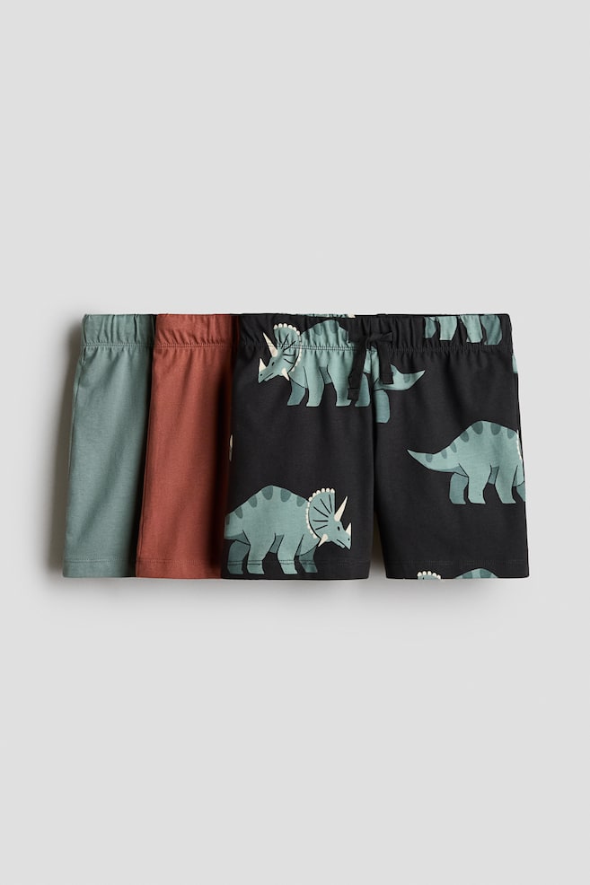 A three-pack of cotton jersey shorts is displaying a solid sage green pair, a solid rust-red pair, and a black pair with a light green triceratops pattern. The shorts are facing front and showing elasticated waistbands, with the frontmost pair featuring a small drawstring tie at the center. - 1