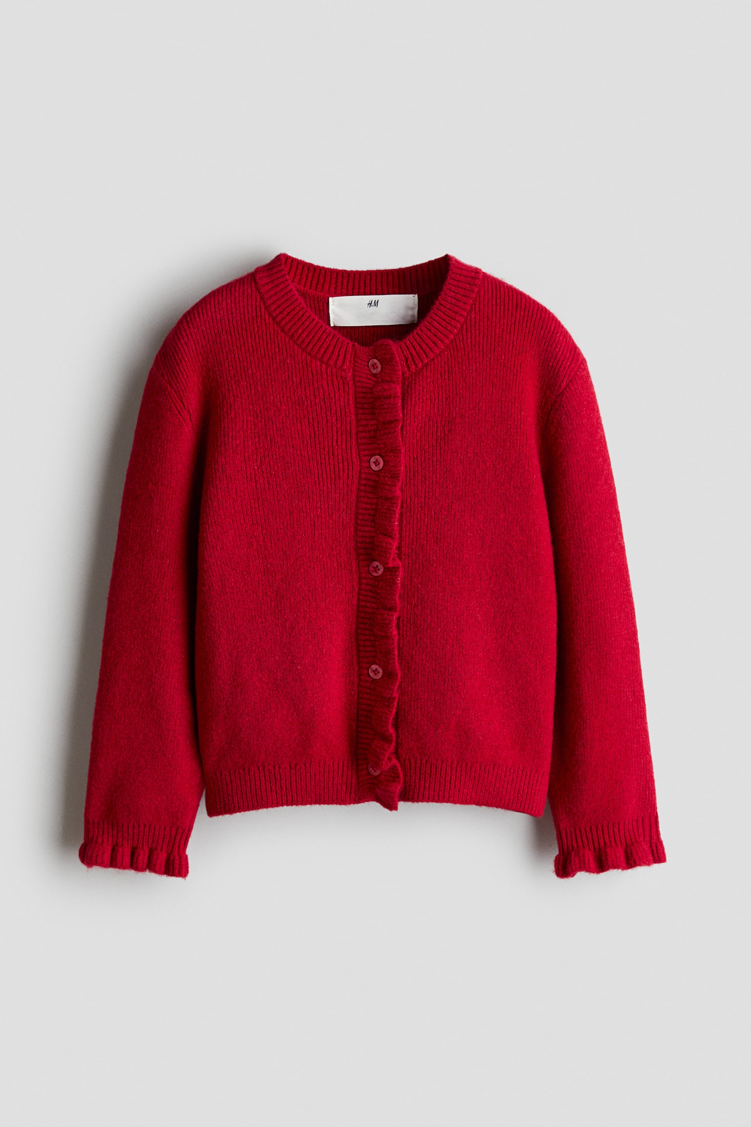 Eine rote Strickjacke ist von vorne zu sehen und prÃ¤sentiert einen runden, gerippt eingefassten Ausschnitt sowie einen Knopfverschluss mit passenden roten KnÃ¶pfen. Sie verfÃ¼gt Ã¼ber feine, gerippte Volants entlang der vorderen Knopfleiste und der ÃrmelbÃ¼ndchen.