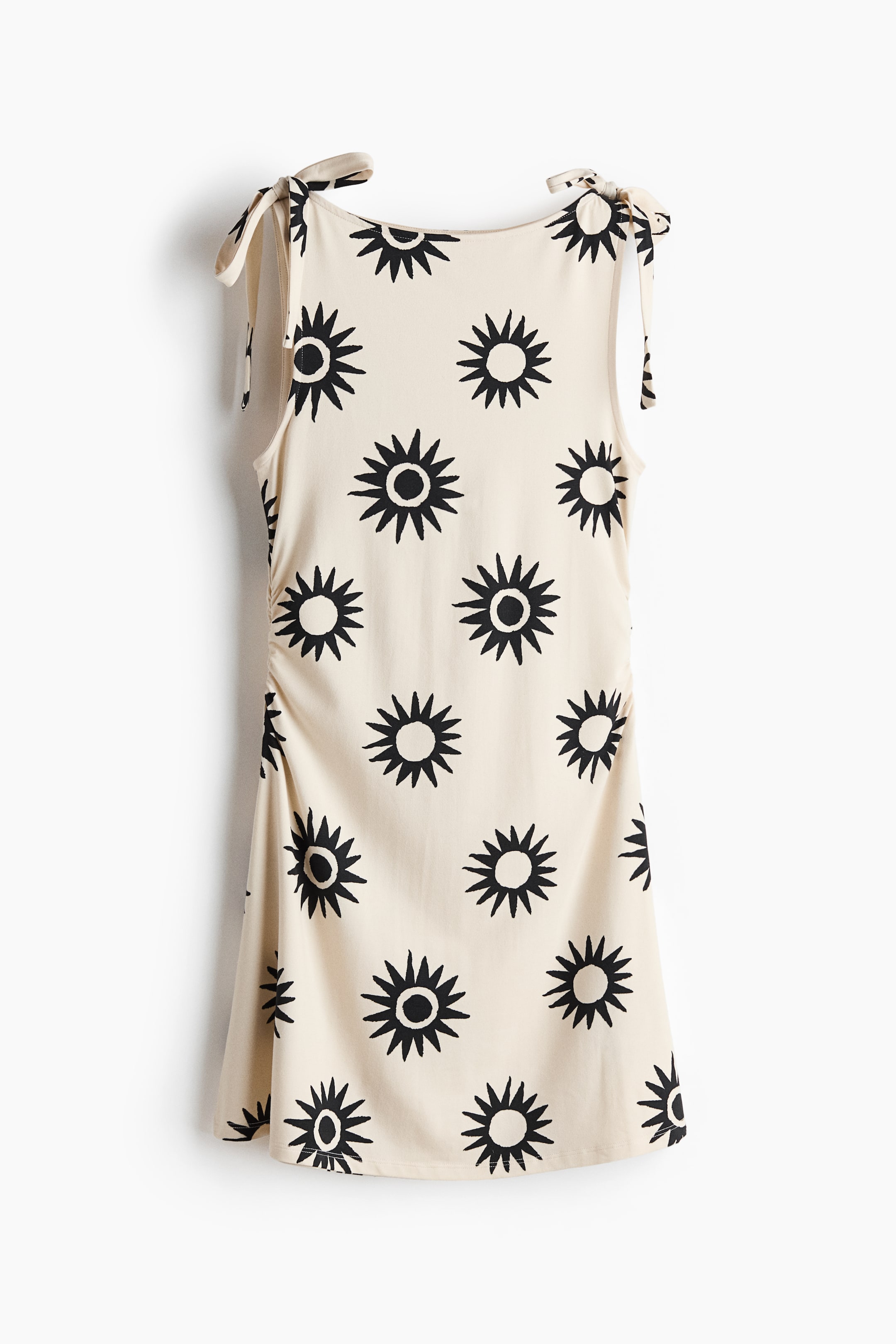 View larger image: The mini dress, front-facing, features a pattern of black suns against a cream backdrop. It boasts a boat neckline, tie-top shoulder straps, gathered detailing at the waist sides, and a subtly A-line skirt.