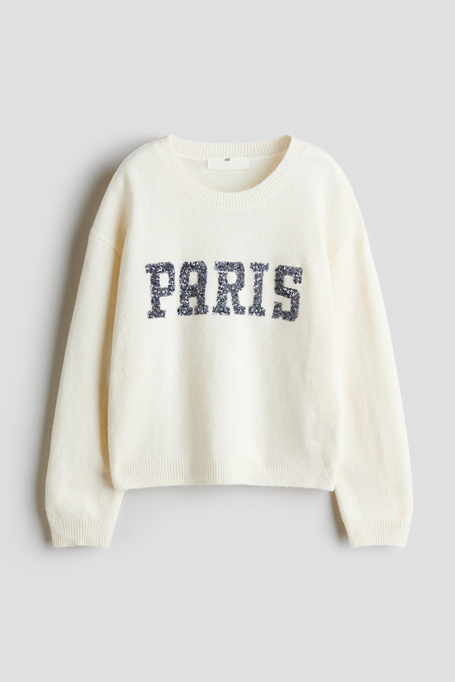 This front view of a loose-fit, off-white knit jumper showcases a round, rib-trimmed neckline, dropped shoulders, and ribbing at the cuffs and hem. The word "PARIS" is spelled out across the chest in dark, sparkling sequins.