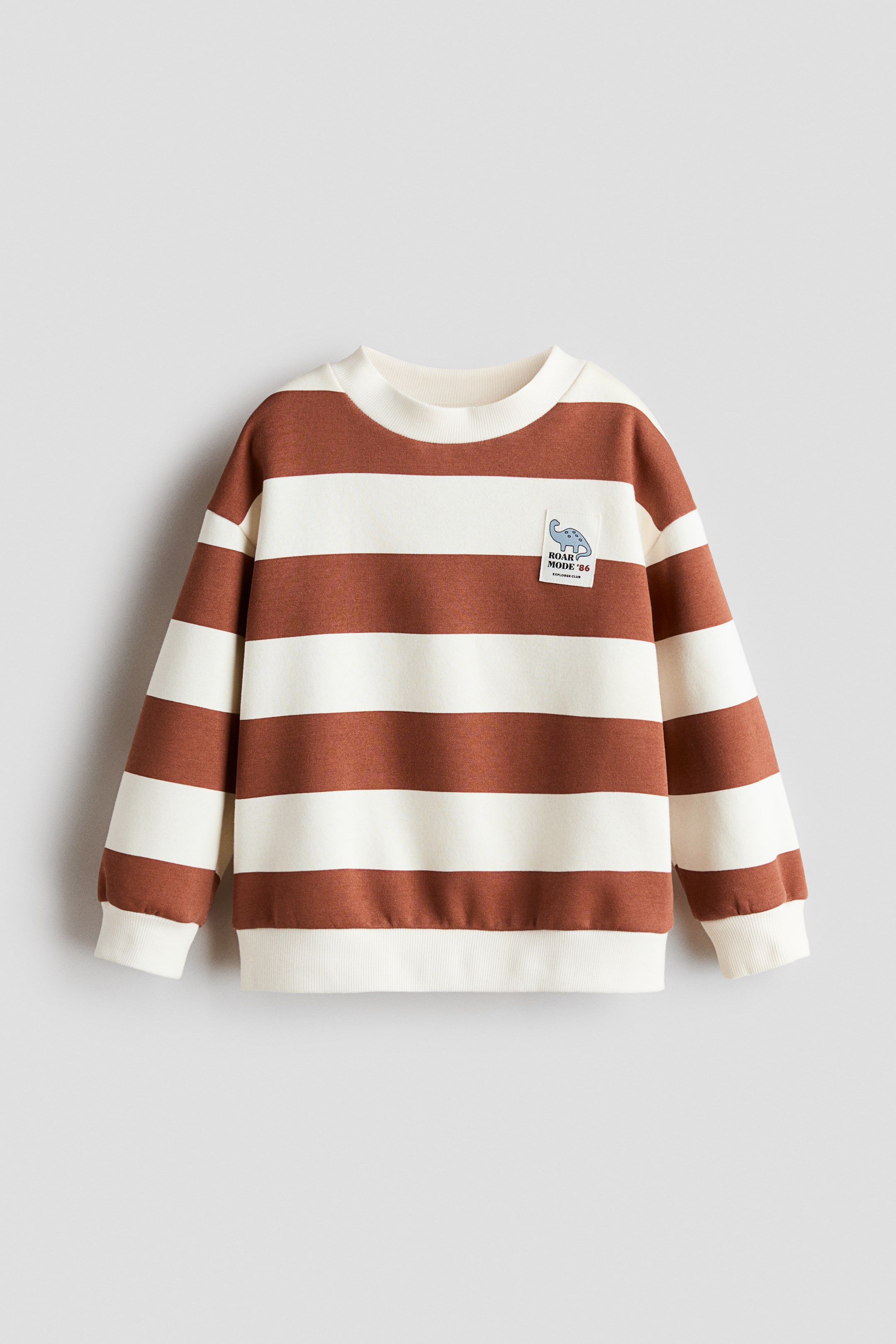 View larger image: A loose-fit sweatshirt is shown from the front, featuring broad horizontal stripes in brown and off-white. The garment has dropped shoulders, a round ribbed neckline, ribbed cuffs, and a ribbed hem, with a small square patch on the upper left chest featuring a dinosaur graphic.