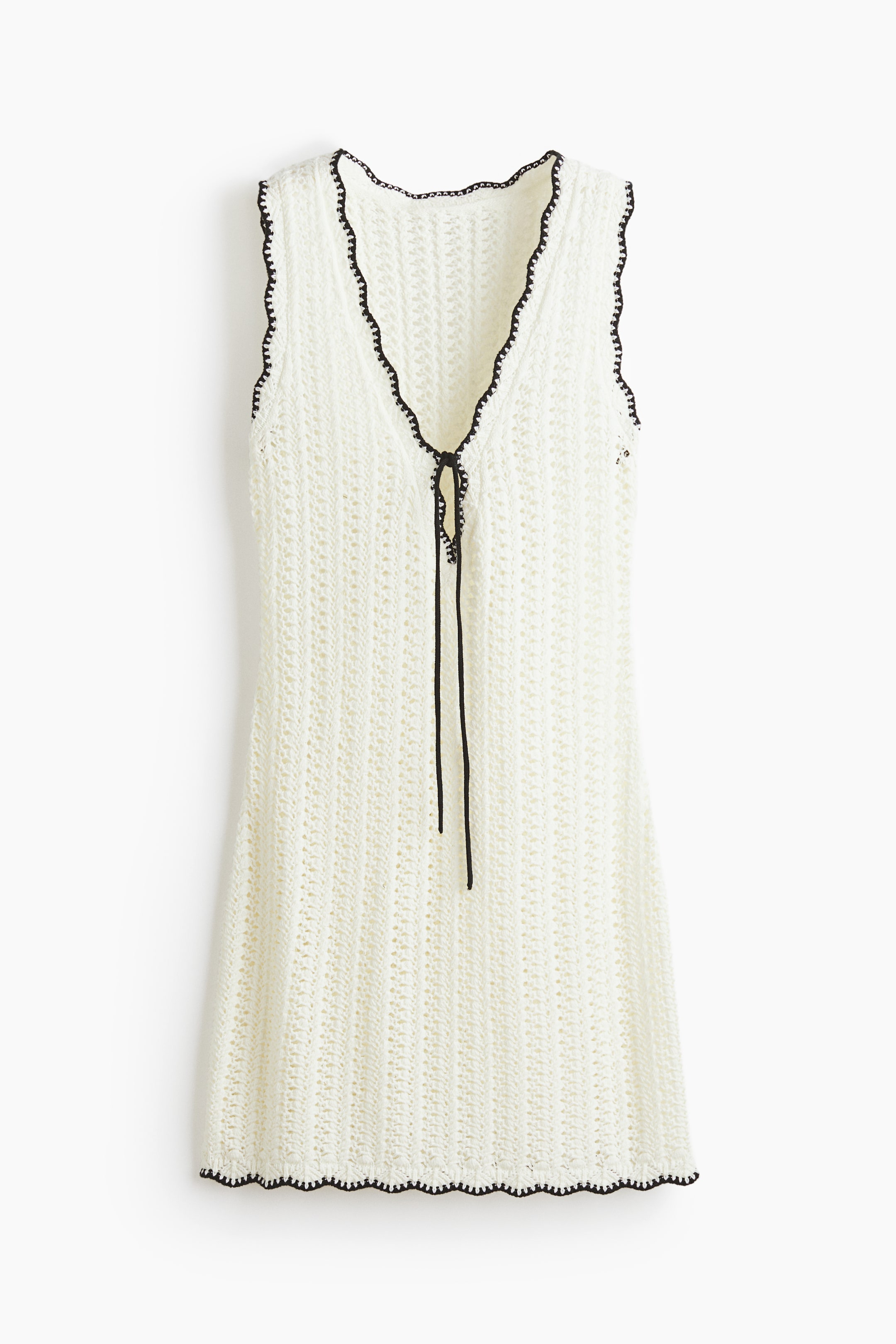 View larger image: An off-white, open-knit crochet dress is hanging front-facing, featuring a V-neckline and sleeveless cut. Black scalloped trim is outlining the neckline, armholes, and hem, while a black drawstring is tying at the front V-neck.