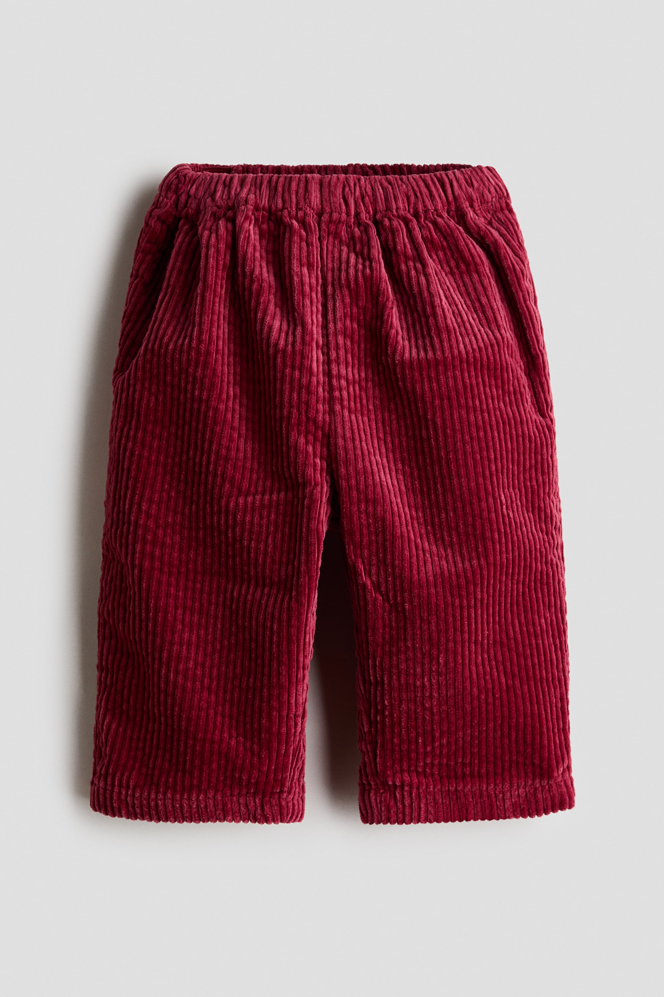 View larger image: A pair of dark red corduroy trousers is laid flat, front facing, showcasing a gathered elasticated waistband, side pockets and a straight-leg cut. The textured, ribbed fabric runs vertically down the length of the legs to the straight hems.