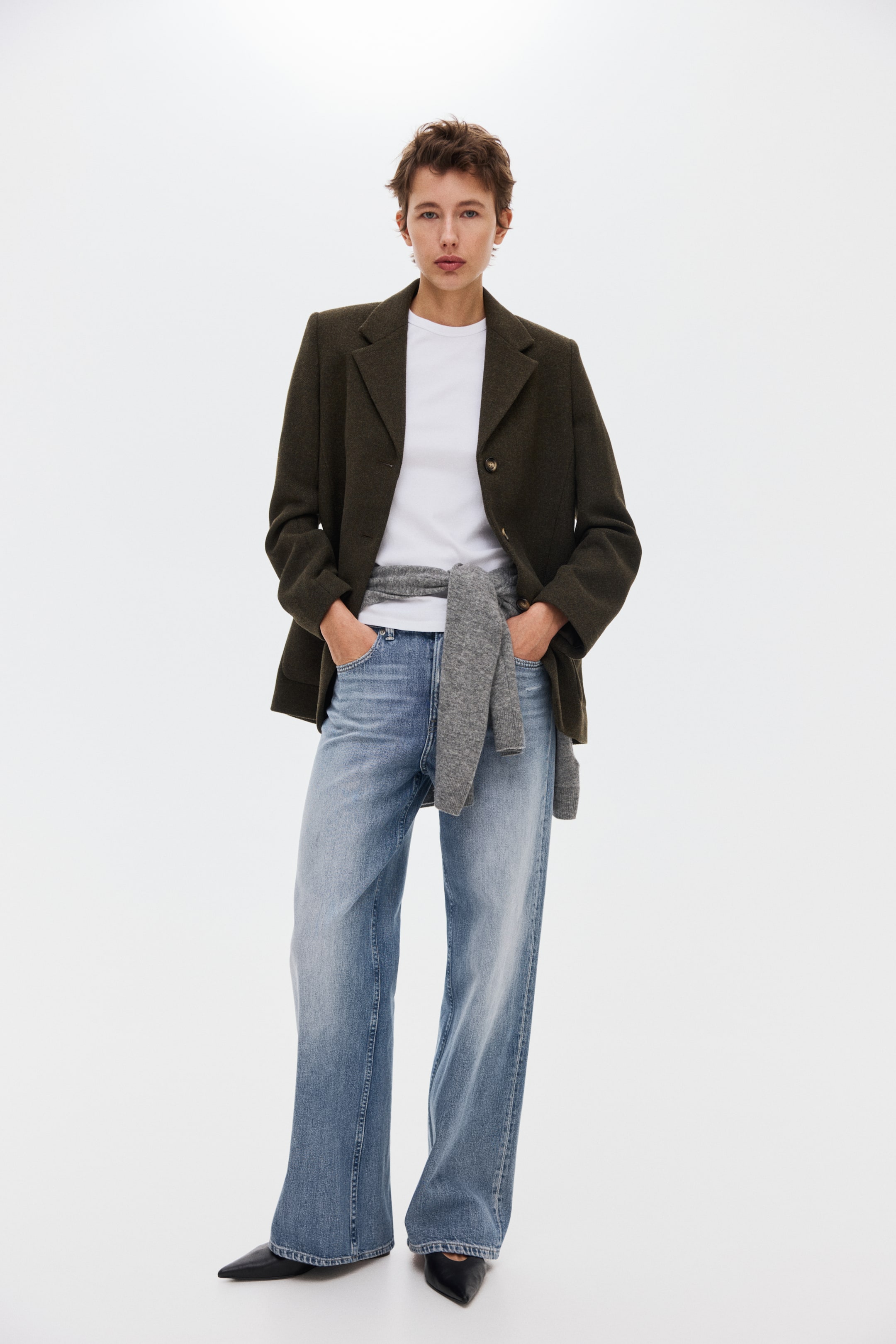 Προβολή μεγαλύτερης εικόνας: Ένα σκούρο ελαιόπρασινο σακάκι, με γιακά με εγκοπή και κλείσιμο με δύο κουμπιά, φοριέται πάνω από ένα λευκό T-shirt με στρογγυλή λαιμόκοψη, ενώ ένα γκρι πουλόβερ είναι δεμένο στη μέση. Ανοιχτό μπλε τζιν με φαρδιά γραμμή συνδυάζεται με μαύρα flats με μυτερή μύτη.