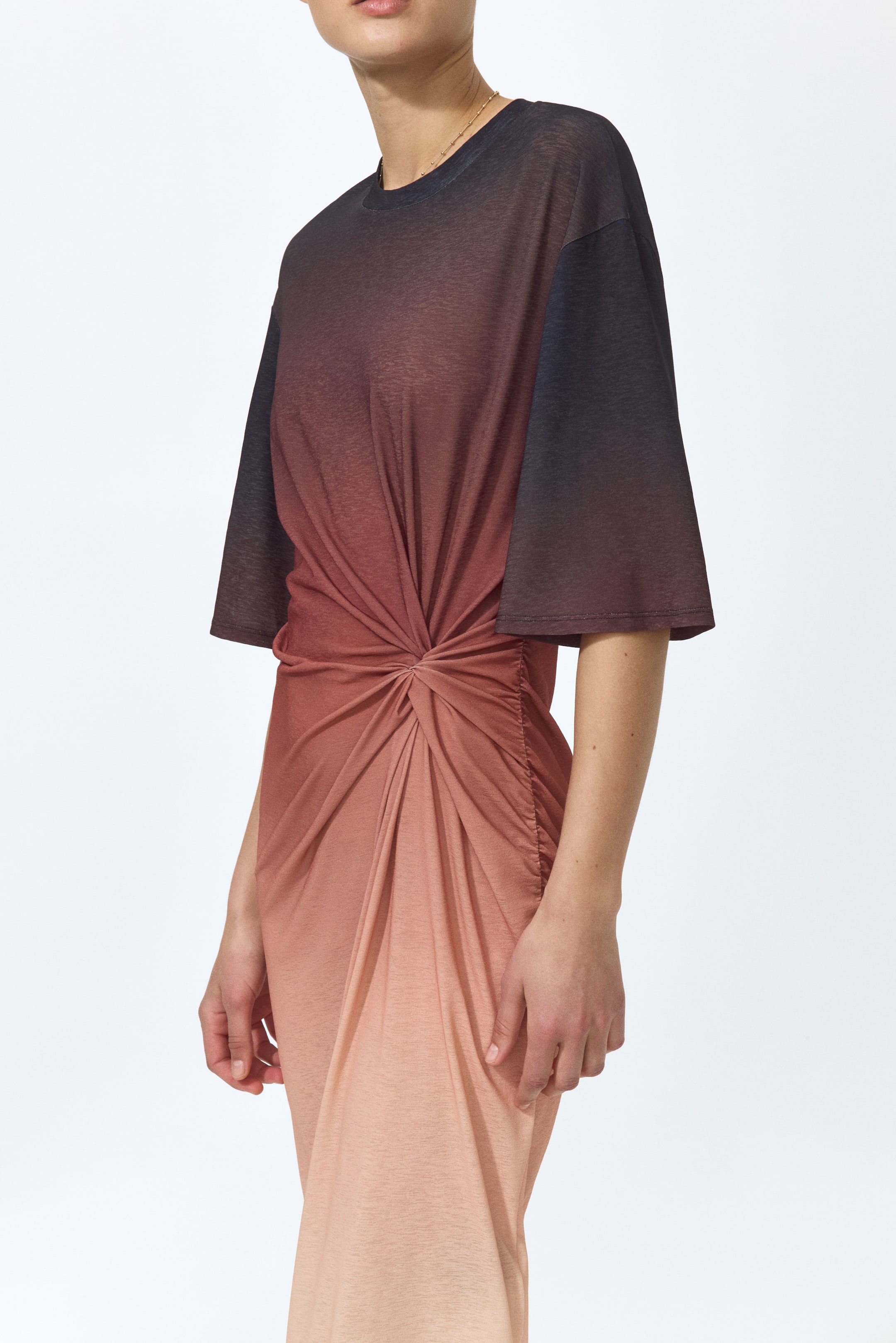 Ampliar la imagen: Un vestido largo de jersey, con un diseño ombré que transiciona de marrón oscuro a melocotón claro, es llevado por la modelo. El vestido cuenta con un escote redondo, hombros caídos, mangas hasta el codo, costuras laterales fruncidas, un detalle de nudo en la cintura y un dobladillo asimétrico.