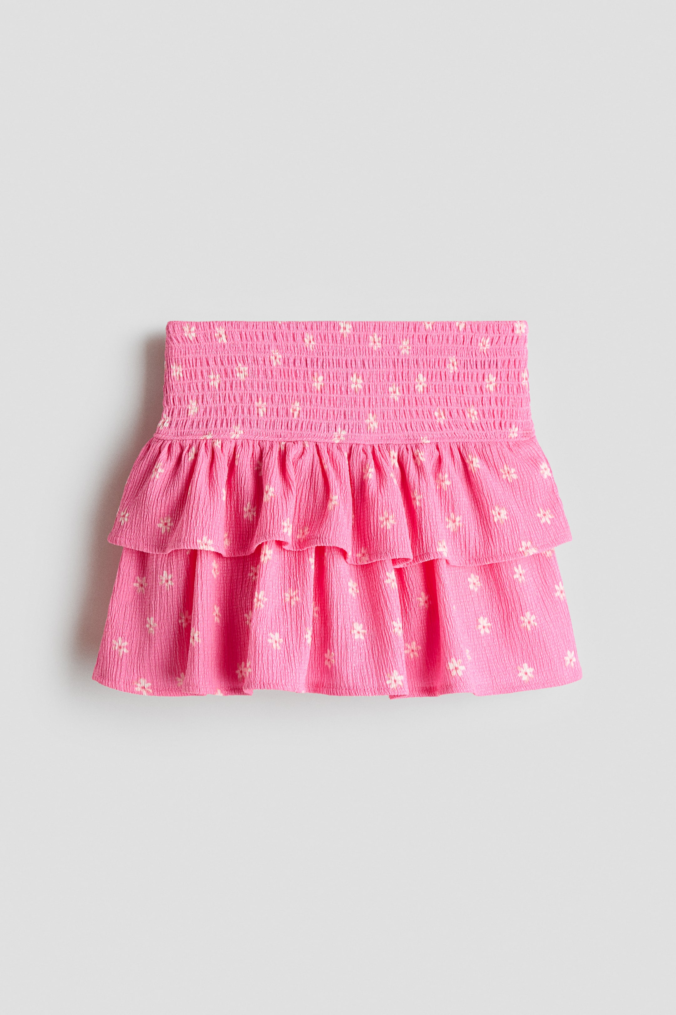 View larger image: A vibrant pink tiered skirt in crinkled jersey is showcased in its front view, highlighting a wide, smocked waistband. Two ruffled tiers shape the skirt's body, adorned with small, light-coloured floral motifs.