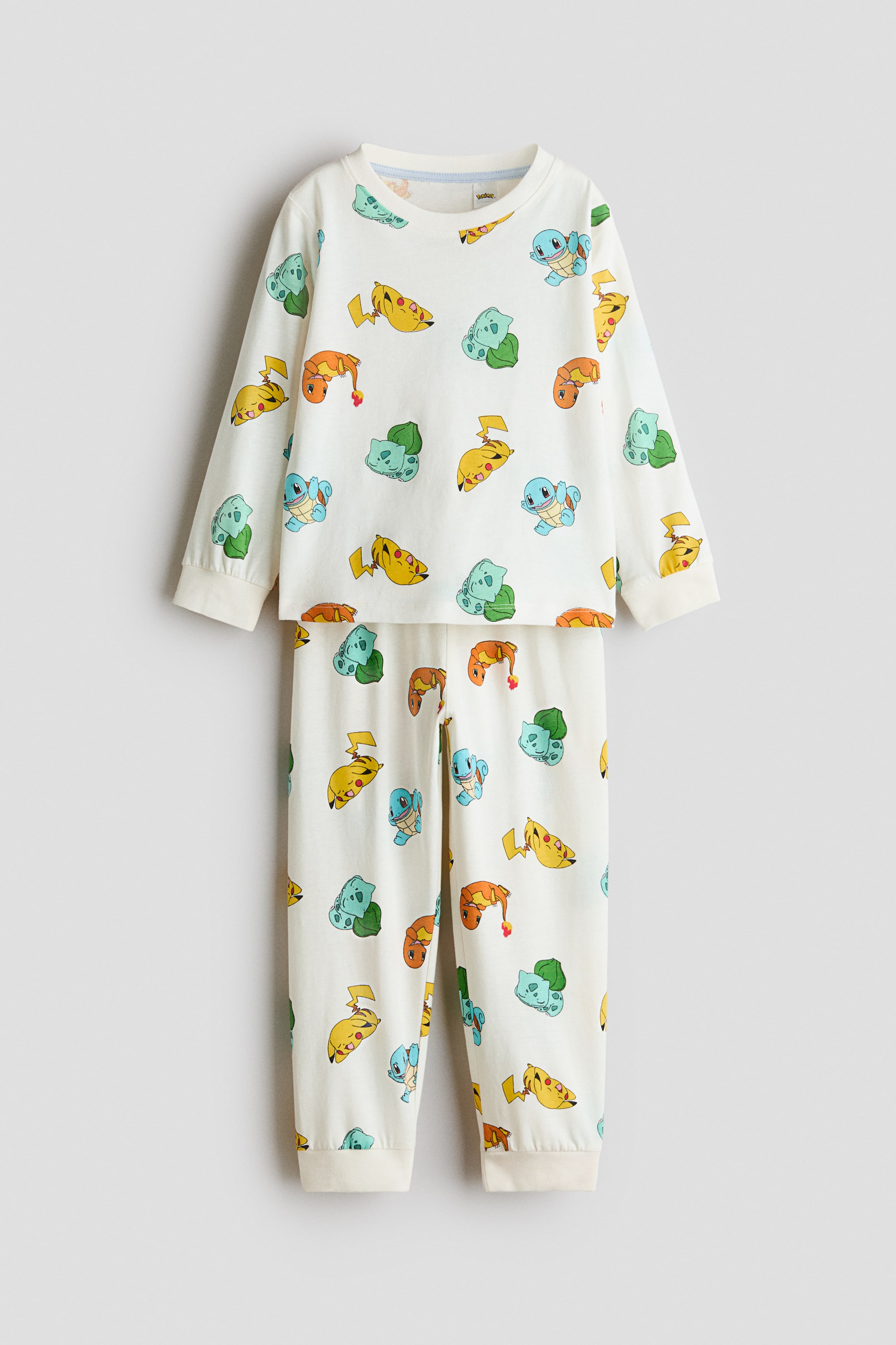 Agrandir l'image: Un ensemble de pyjama en jersey de coton couleur crème, présenté de face, affiche un imprimé intégral de personnages Pokémon. Le haut à manches longues présente un col rond côtelé et des poignets côtelés, tandis que le pantalon assorti arbore des bords-côtes aux ourlets.