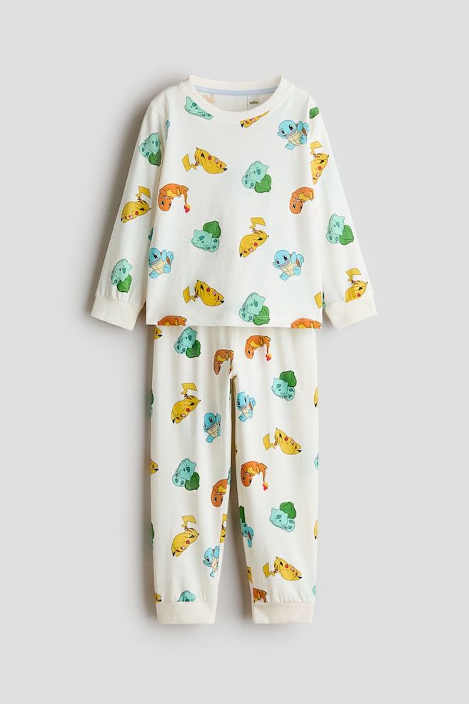Et cremefarvet pyjamassÃ¦t i bomuldsjersey ses forfra med et all-over print af Pokï¿½mon-figurer. Den langÃ¦rmede overdel har en rund, ribbet halsudskÃ¦ring og ribbede manchetter, mens de matchende bukser har ribbede manchetter ved buksebenene. - 1
