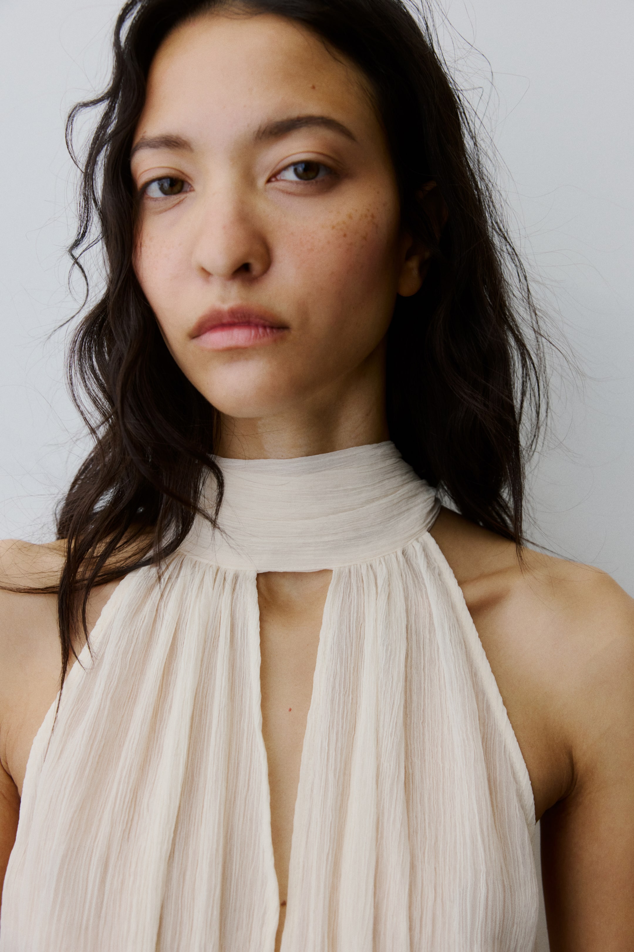 Ampliar la imagen: Un top sin mangas de color beige claro presenta un cuello alto fruncido y un corte de ojo de cerradura central, con la tela arrugada cayendo suavemente.