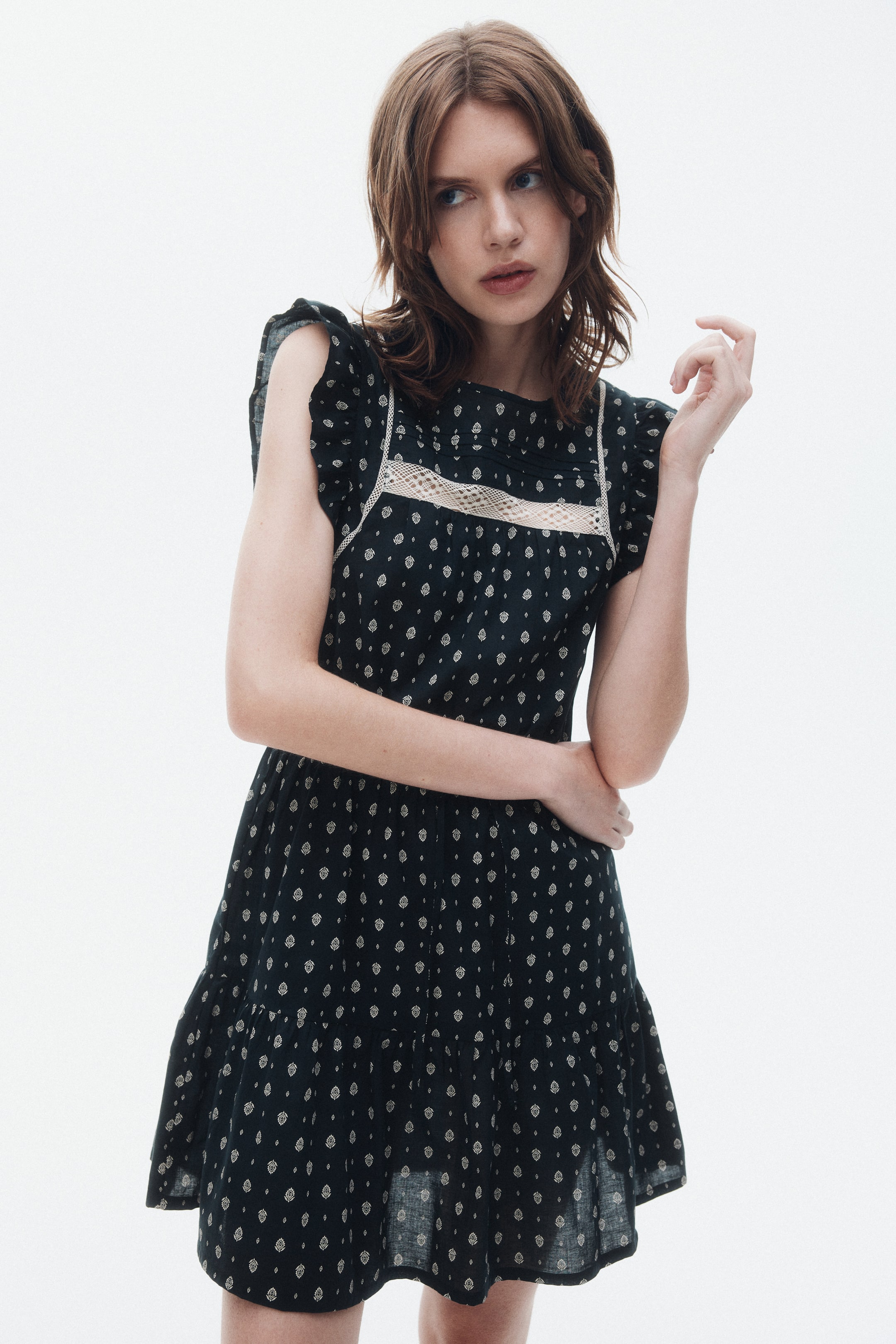 Xem ảnh lớn hơn: Một chiếc váy mini màu tối với họa tiết nhỏ, màu sáng lặp lại, cổ tròn và ống tay cánh rớt. Chiếc váy này có điểm nhấn là một lớp vải ren ngang chạy ngang qua phần trên ngực, được viền bởi các đường viền trắng thẳng đứng và chân váy tầng với đường viền nhún, được nhìn từ góc độ hơi nghiêng phía trước.