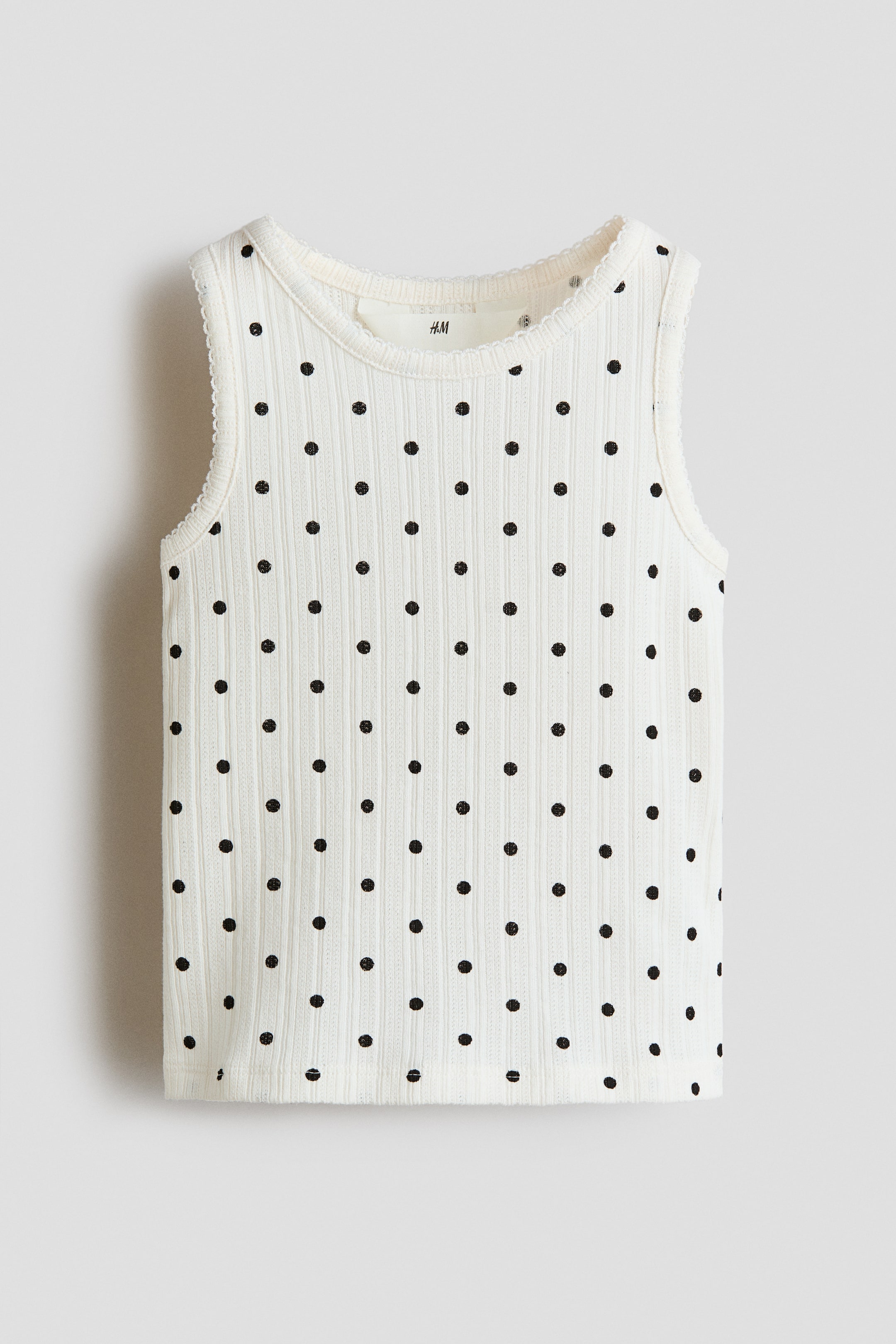 View larger image: The white ribbed tank top showcases a pattern of black polka dots, presented front-facing. It features a round neckline and scalloped trim along the neck and armholes.