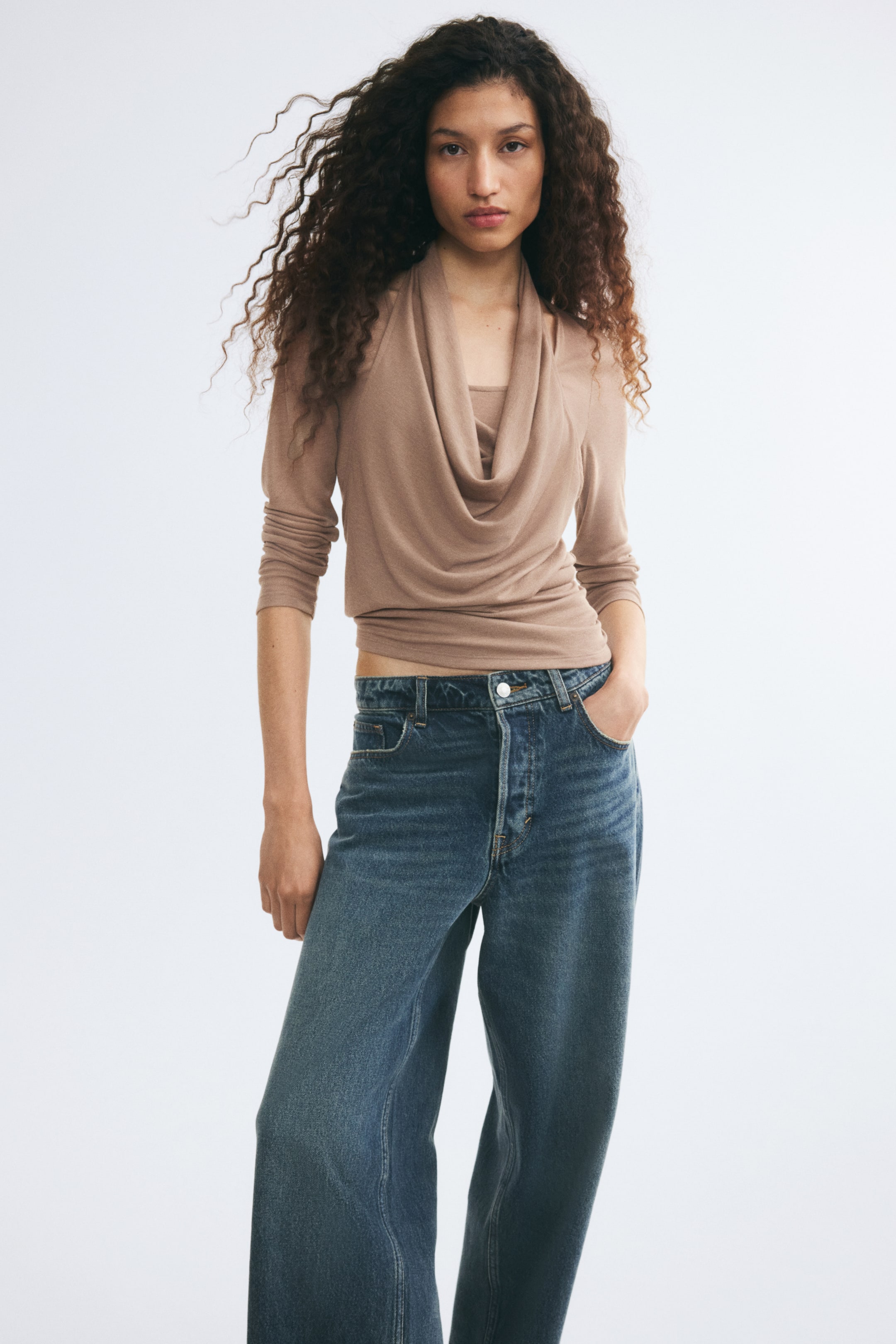 Agrandir l'image: Un haut en maille beige avec un col bénitier drapé et des manches longues est porté par le mannequin. Le haut est associé à un jean en denim bleu à jambes larges, le mannequin ayant une main glissée dans une poche avant.