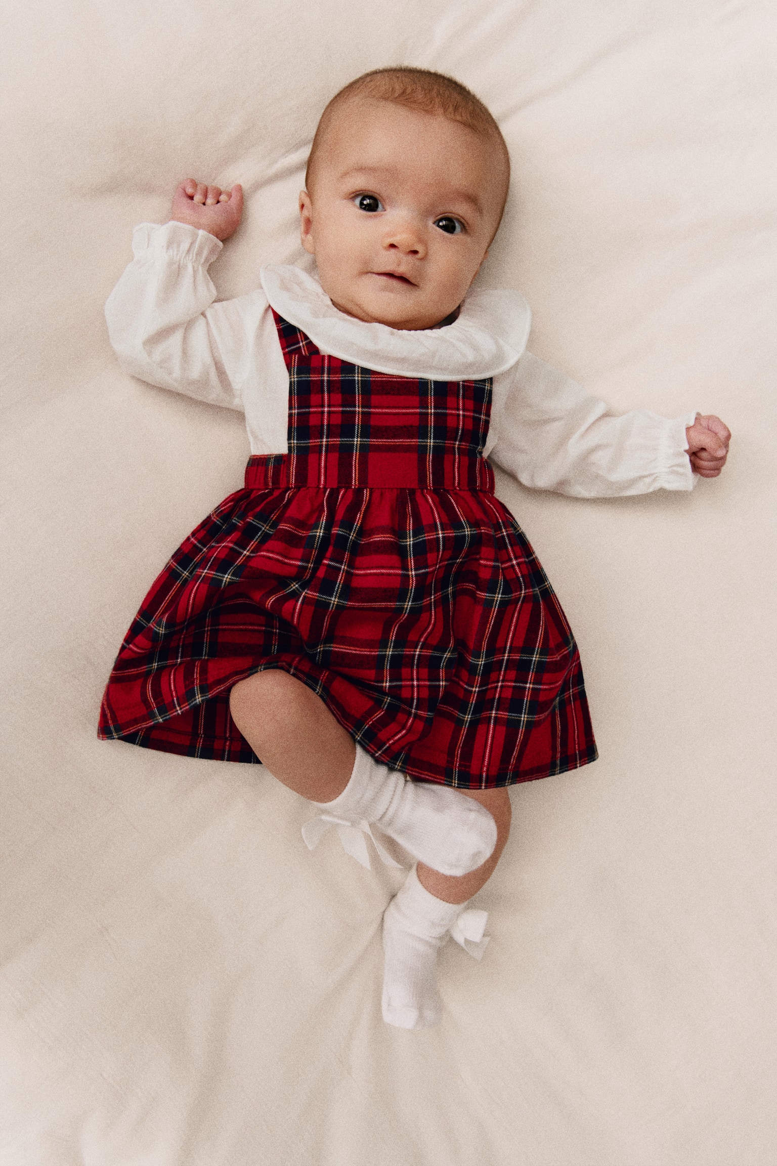 A baby’s two-piece set is shown from the front, consisting of a red tartan plaid pinafore dress layered over a white long-sleeved bodysuit. The dress features a square bib and a gathered flared skirt, while the bodysuit has a wide frill collar and ruffled cuffs. - 1