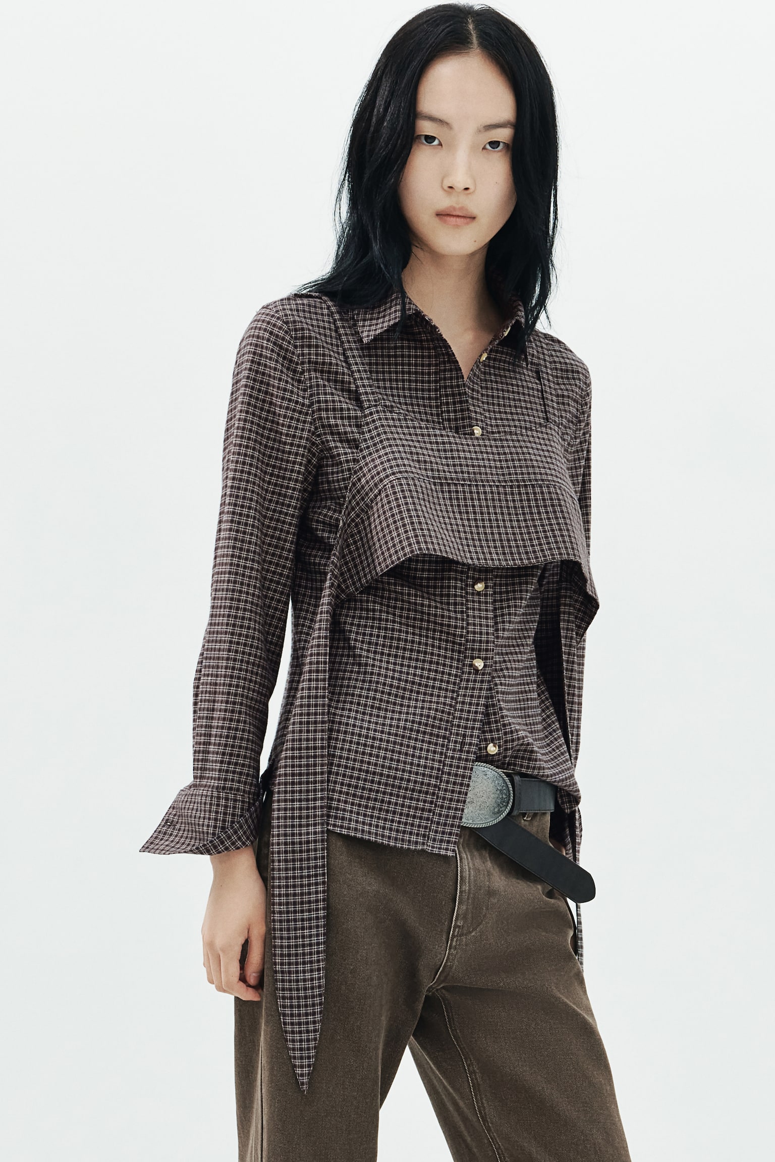 A plaid cotton twill shirt is shown from the front, featuring a classic collar, a button-down front with metallic buttons, and long sleeves with wide cuffs. A matching plaid crop top layer with narrow shoulder straps is layered over the chest of the shirt, creating a double-layered look. - 1