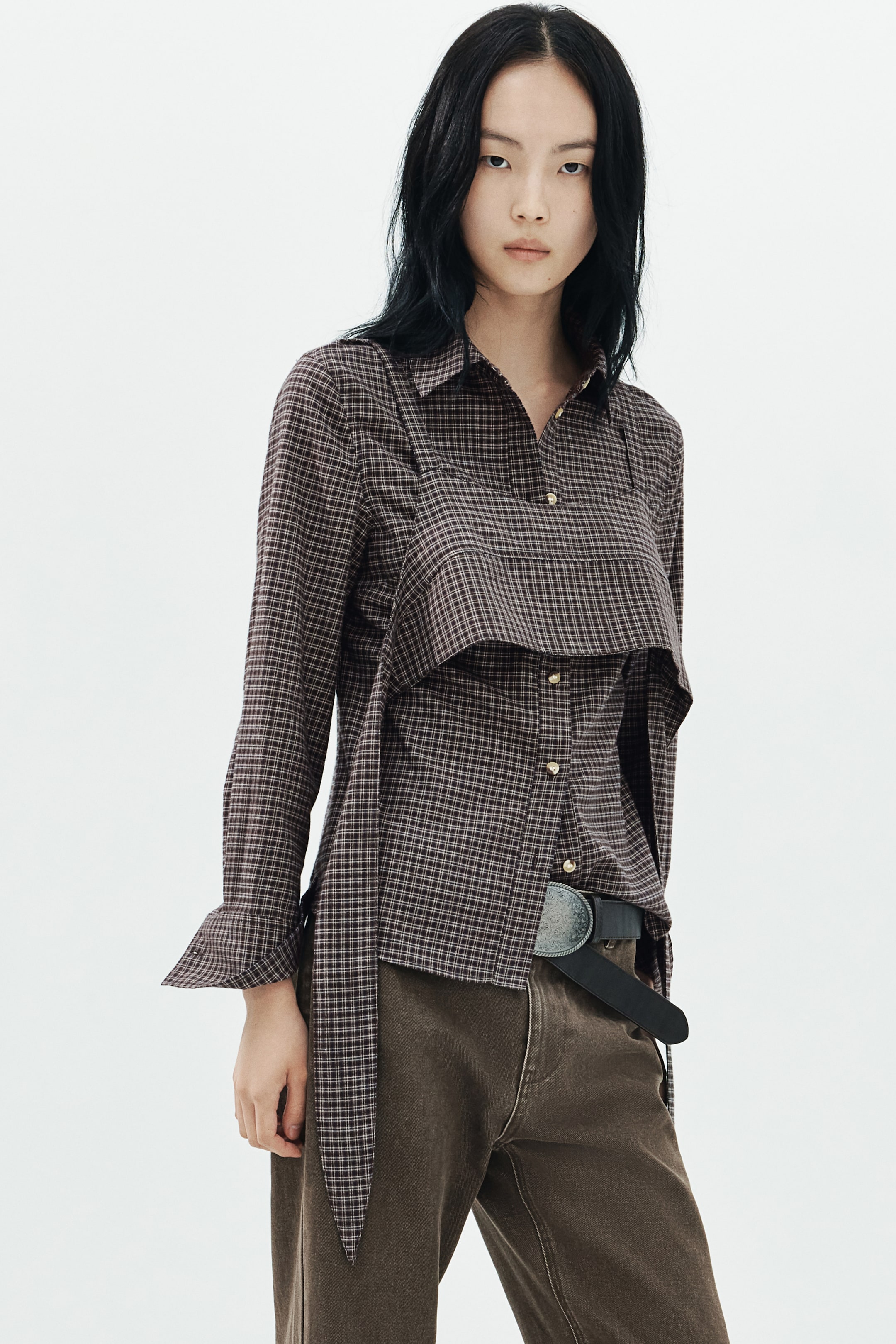 View larger image: A plaid cotton twill shirt with a collar, a button-down front, and long sleeves is being worn by the model. The shirt features an integrated matching crop top layer with narrow shoulder straps and long, wide ties hanging at the side, styled with brown trousers.