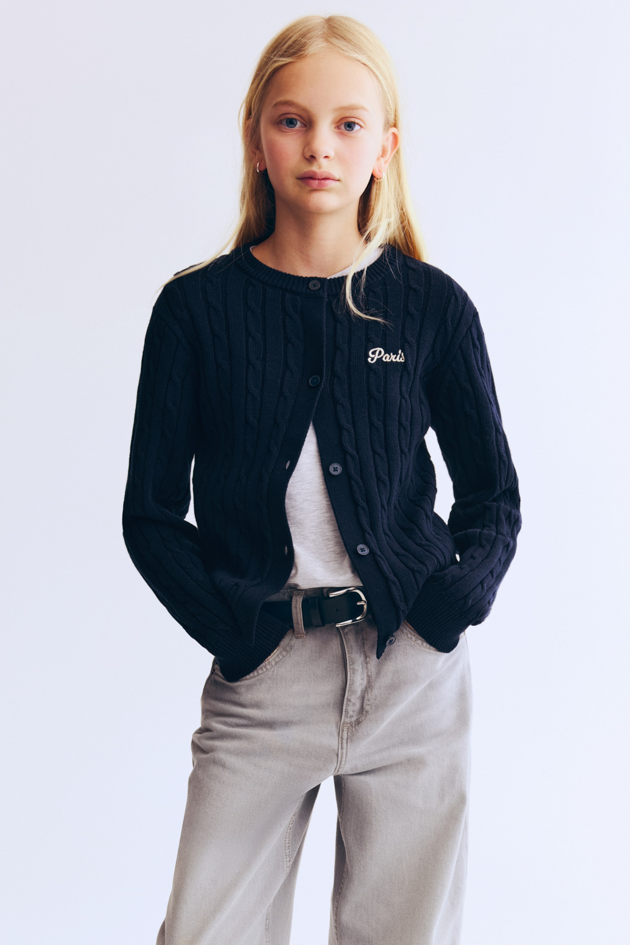 View larger image: A navy-blue cable-knit cardigan is worn open over a white T-shirt. It features a round neck, a button-front closure, and white "Paris" embroidery on the left chest, with ribbed cuffs and hem visible.