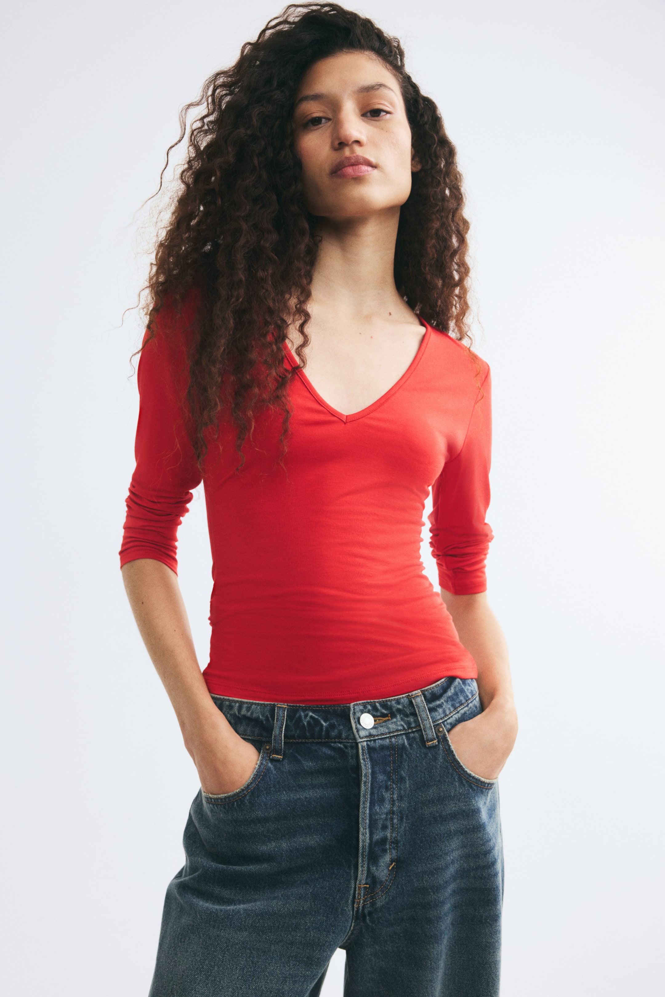 View larger image: A fitted red top with a deep V-neck and 3/4-length sleeves is being worn by the model. The top is styled with blue denim jeans, with the model resting their hands in the front pockets.