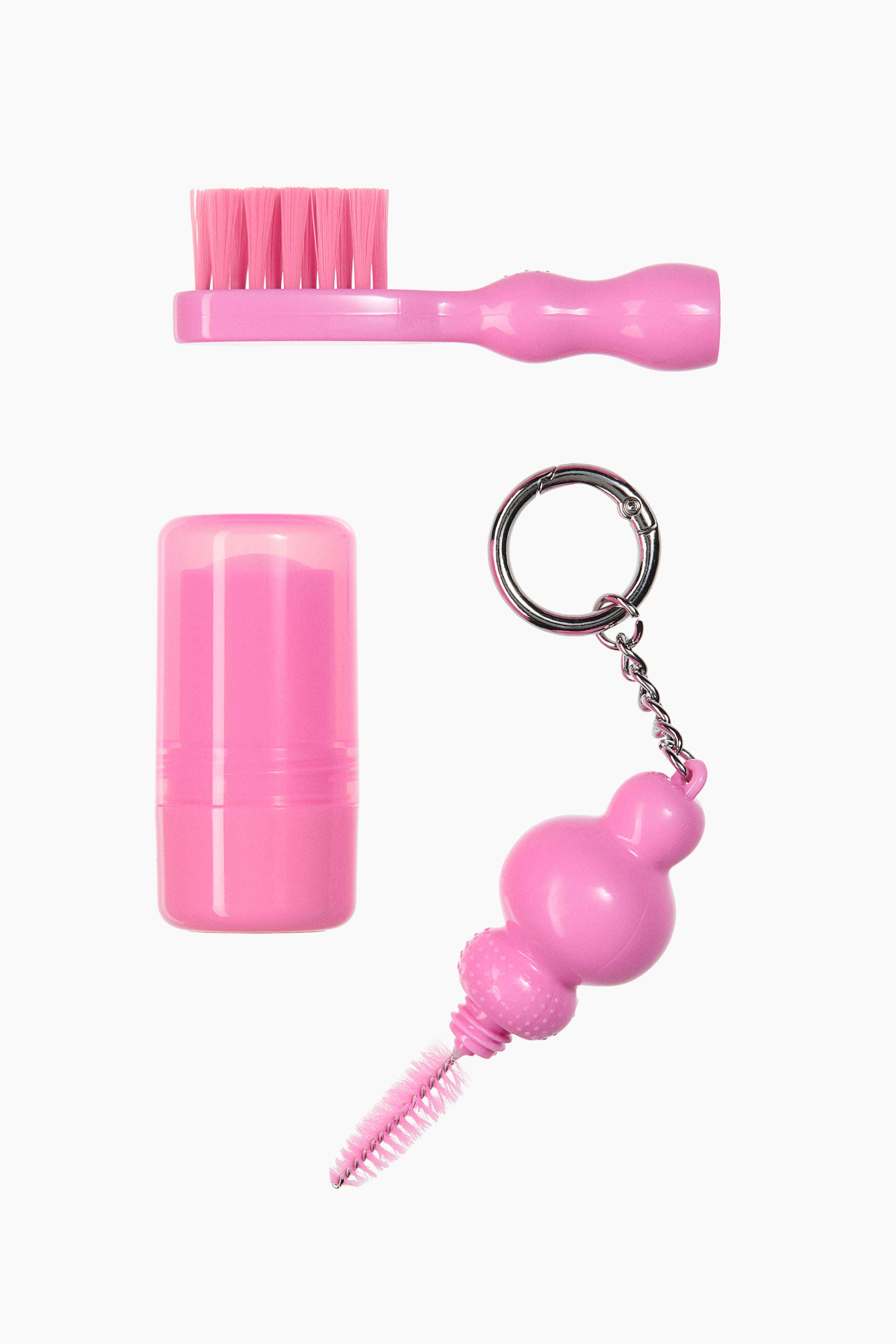 View larger image: A pink hair styling kit displays three separate components: a mini styling brush with pink bristles at the top, a small cylindrical pink wax stick with a translucent cap at the bottom left, and a mini spoolie brush featuring a bulbous pink handle attached to a silver-toned metal keychain clasp at the bottom right.