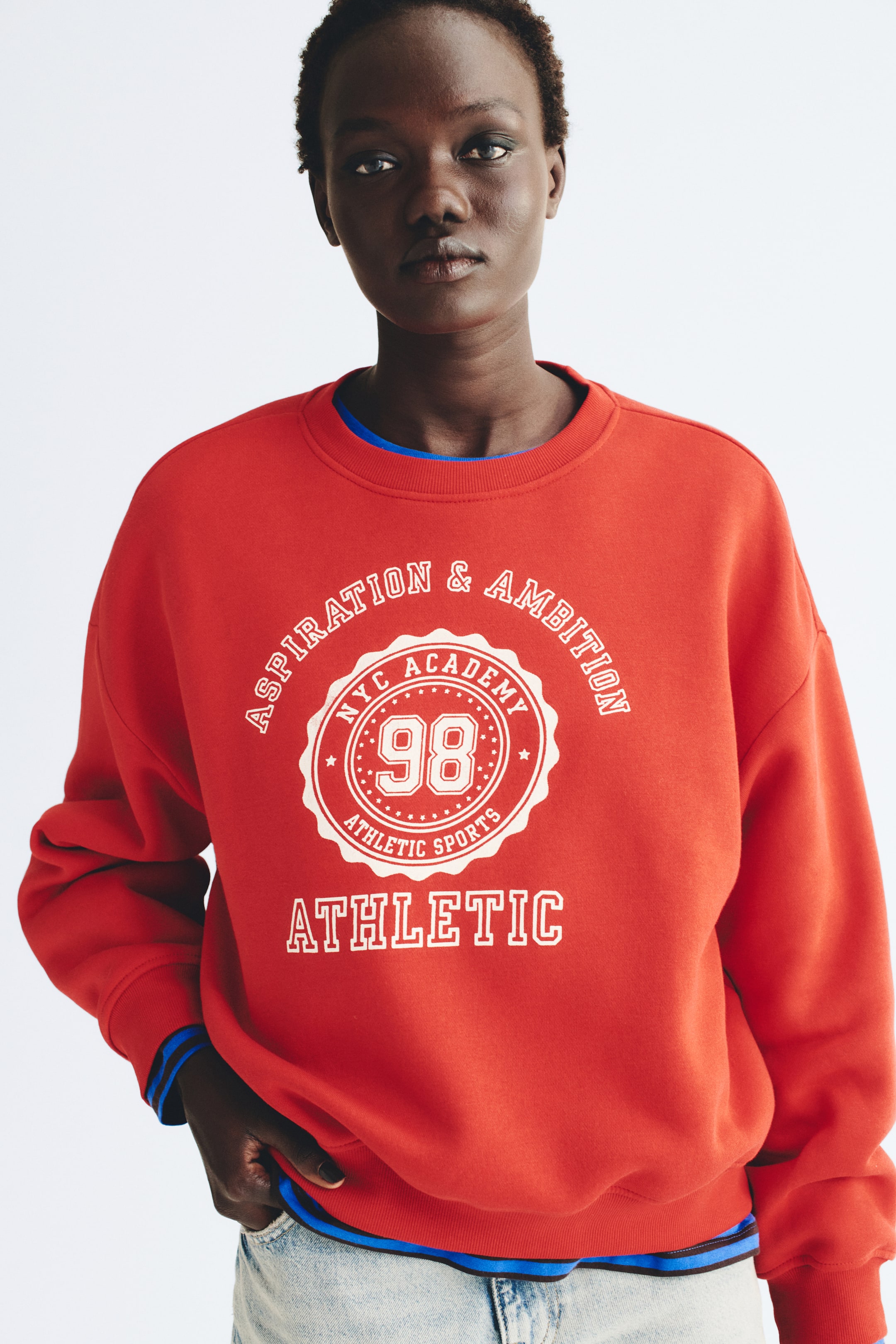 Voir une image plus grande: Un chandail à capuchon surdimensionné rouge, orné d’un imprimé graphique de style universitaire blanc sur la poitrine, est porté par le mannequin. Le chandail à capuchon présente un col rond côtelé, des épaules tombantes ainsi que des poignets et un ourlet côtelés, et il est stylisé par-dessus un haut bleu superposé et un jean en denim délavé pâle.