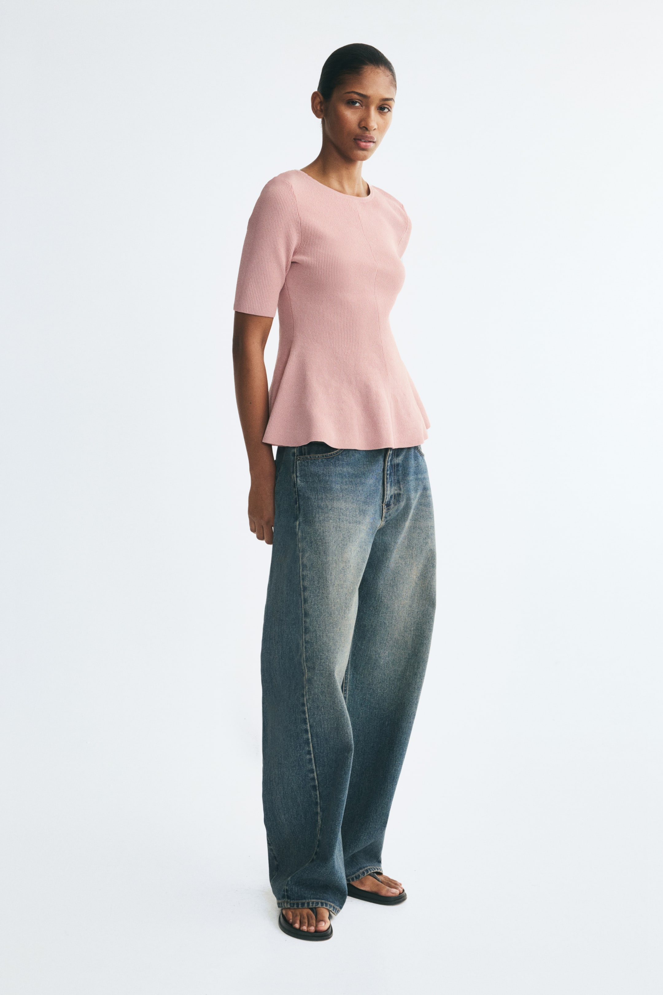 View larger image: The model is wearing a pink knit top featuring a round neckline, elbow-length sleeves, and a flared peplum hem. The top is styled with wide-leg blue jeans.
