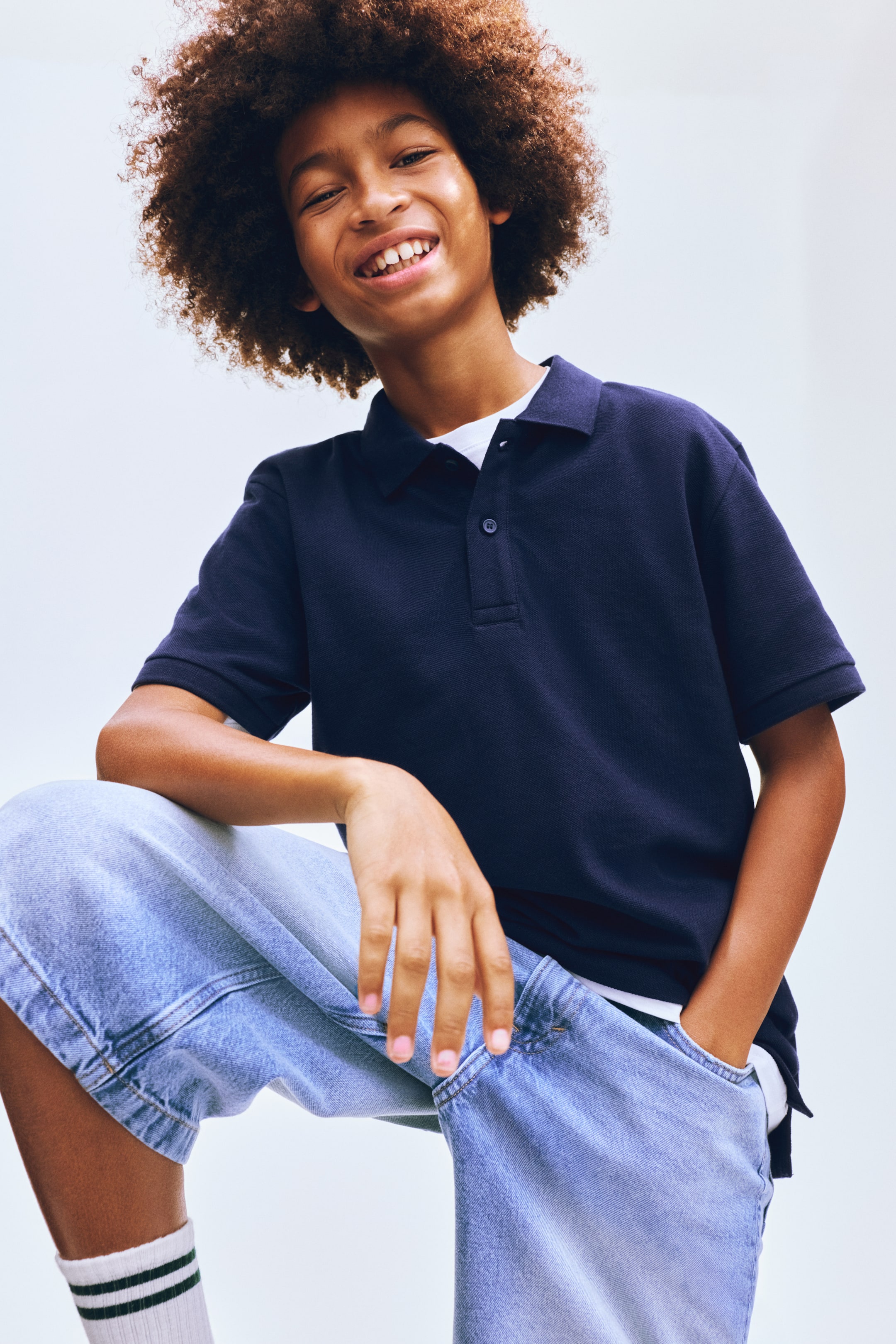 Agrandir l'image: Un polo bleu marine avec un col pointu, une patte de boutonnage à deux boutons et des manches courtes, superposé sur un t-shirt blanc. Le polo est présenté de face, légèrement incliné, et est porté avec un short en denim bleu clair, ample, affichant des détails de couture proéminents et des chaussettes blanches montantes avec deux rayures vertes.
