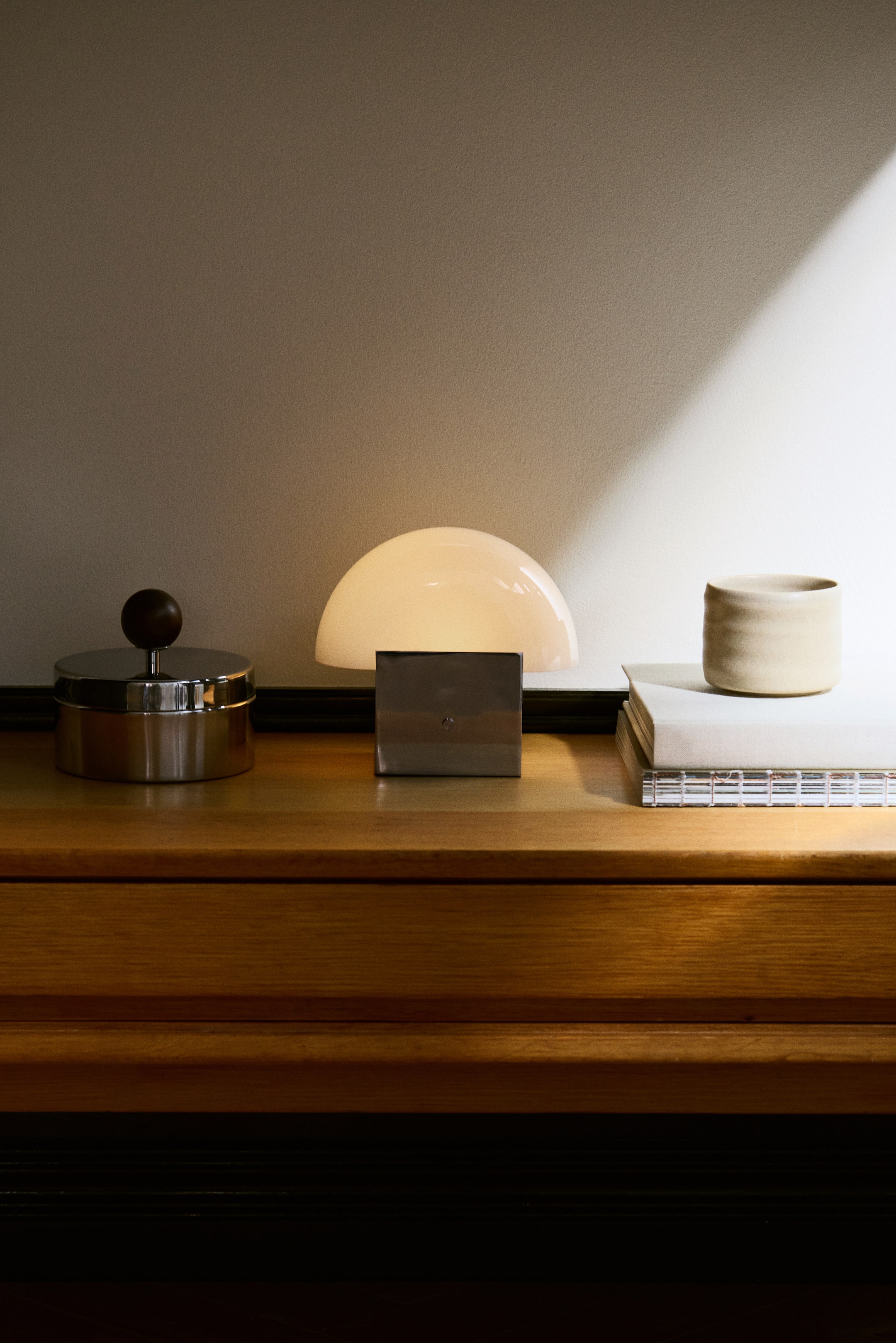 Ver imagen más grande: Una lámpara de mesa recargable está orientada hacia el frente y presenta una pantalla de cristal translúcido en forma de cúpula que emite un resplandor cálido. La pantalla descansa sobre una base rectangular de bloque metálico pulido, mostrando un diseño geométrico minimalista.
