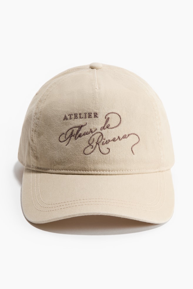 A front-facing beige baseball cap features a six-panel design and a curved brim, with dark brown embroidery on the front displaying 'ATELIER' in block letters above 'Fleur de Rivera' in script. - 1