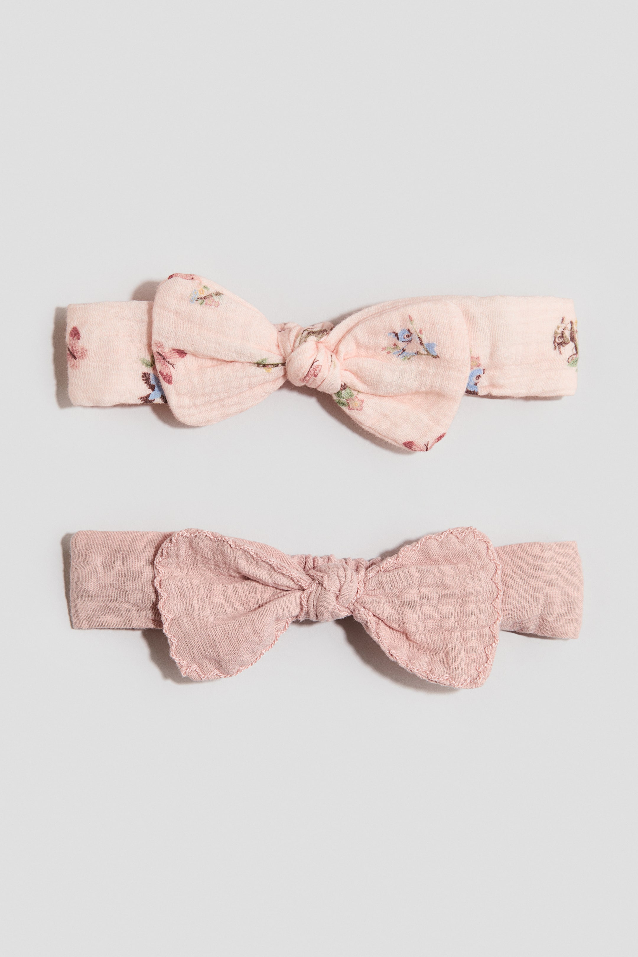 View larger image: Two headbands are displaying their FRONT. The top headband is a light pink gauze, featuring a small floral print and a tied bow, while the bottom headband is a solid pink gauze with a bow featuring scalloped, stitched edges.