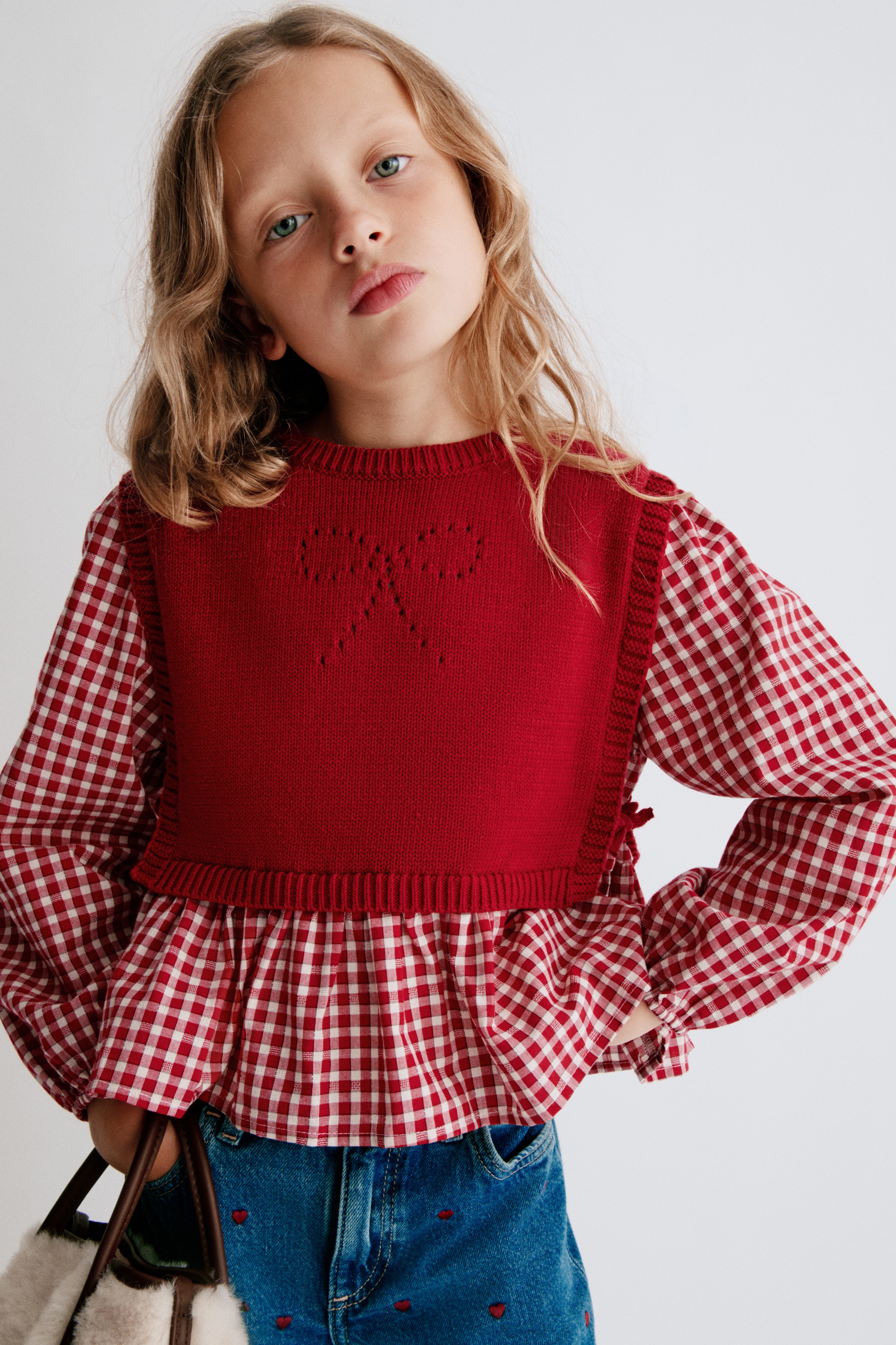 A red 2-in-1 top is displayed with a solid red knit vest layered over a red-and-white gingham shirt, viewed from the front. The knit vest features a crew neck, ribbed trims, and a pointelle bow design at the centre chest, while the shirt underneath showcases long sleeves with elasticated cuffs and a ruffled peplum hem.