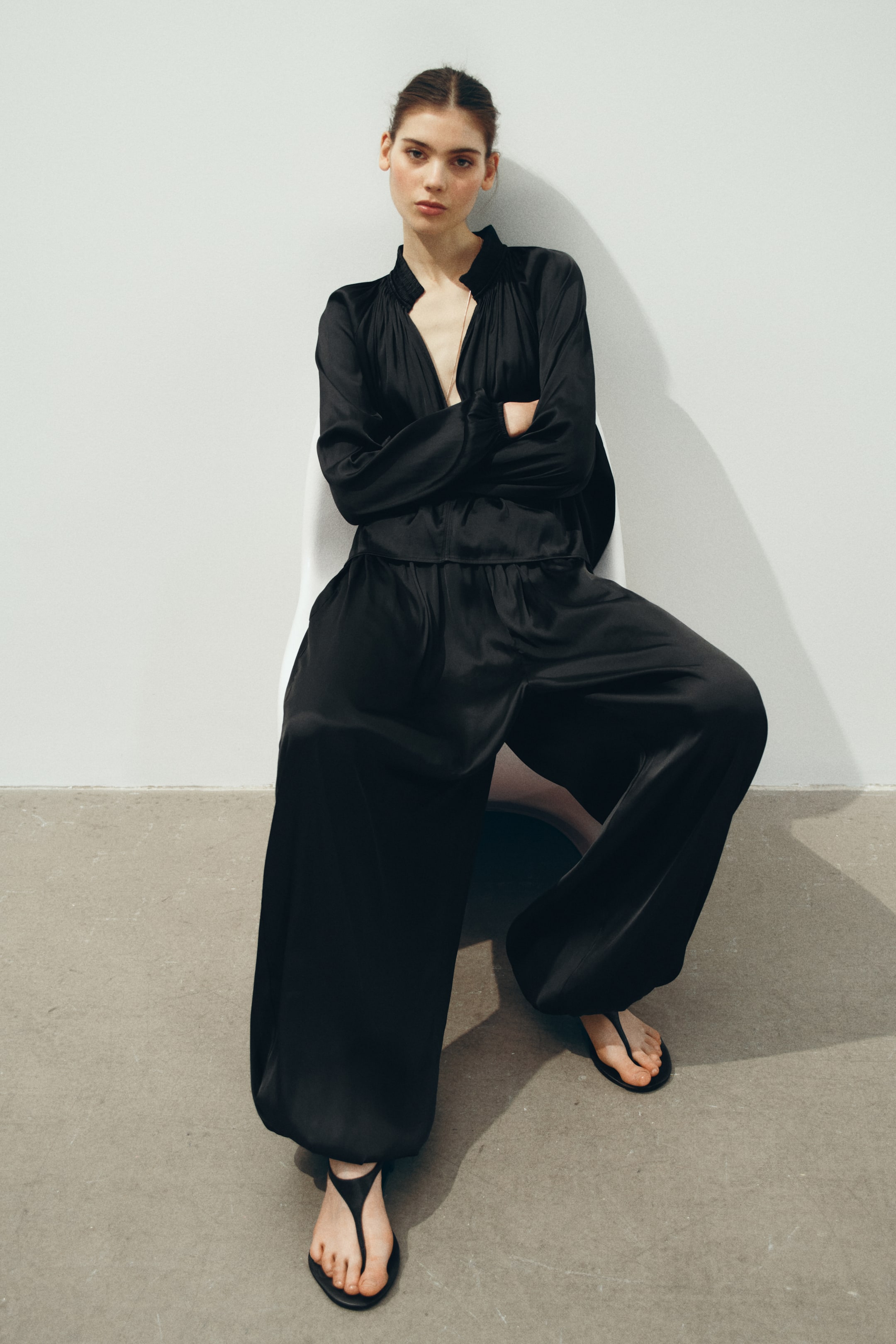 View larger image: A black satin two-piece ensemble featuring a top with a gathered band collar and deep V-neck, long blouson sleeves, and wide-leg trousers with an elasticated waist, flowing to the floor.