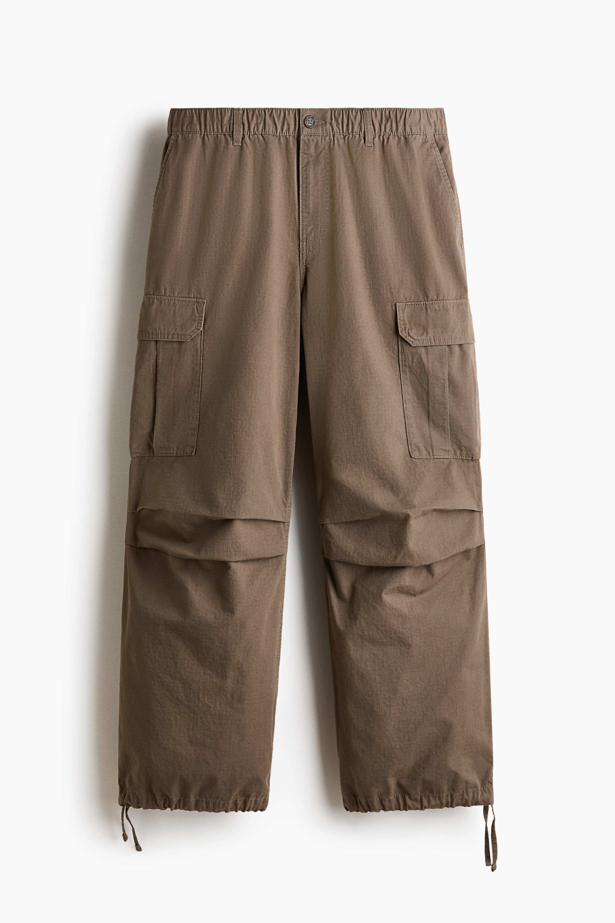 View larger image: Front-facing cargo trousers in cotton twill feature a covered elasticated waist with a drawstring and a fake fly. They are designed with diagonal side pockets, flapped patch leg pockets, pleats at the knees, and drawstrings with cord stoppers at the hems, presenting a loose fit.