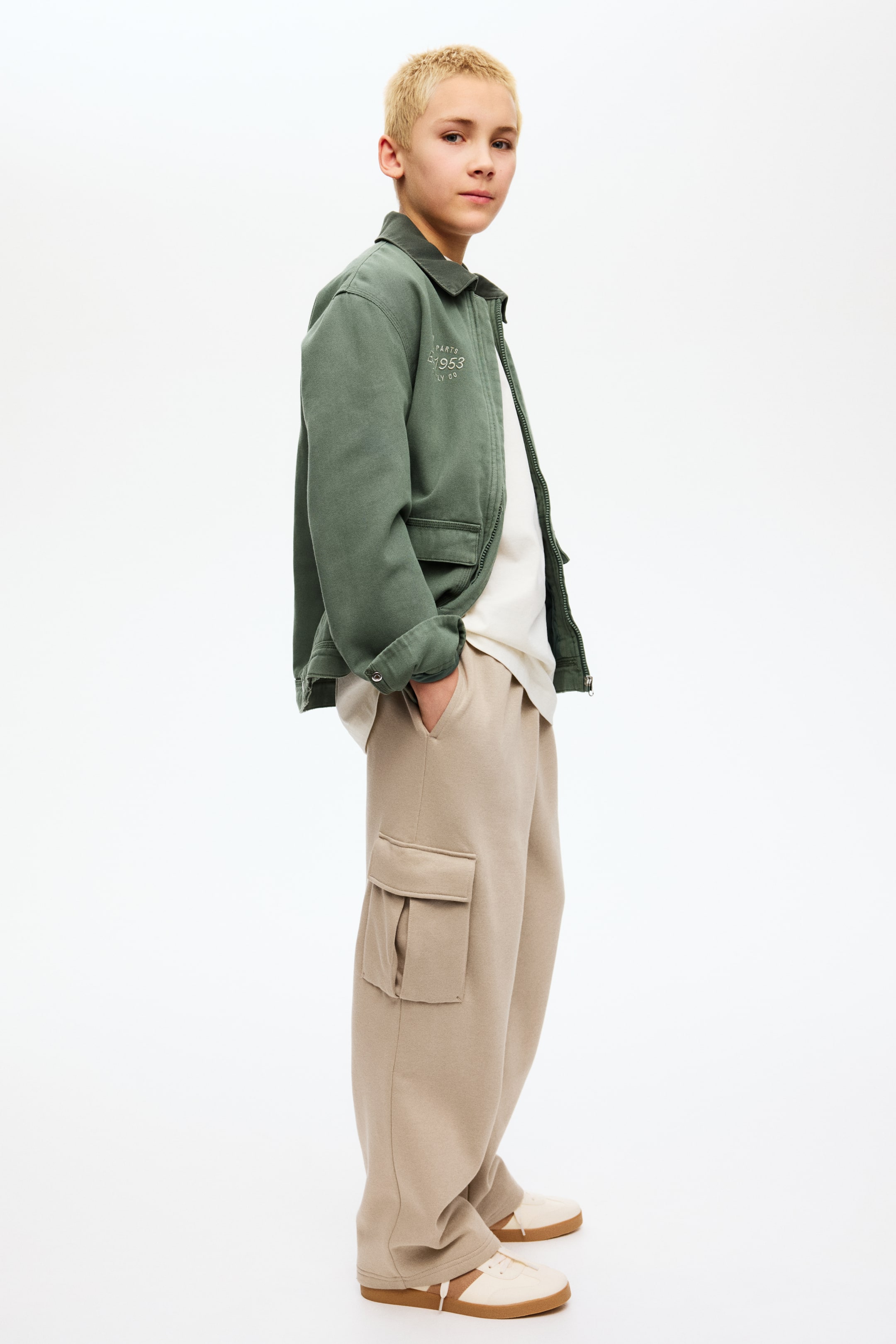 Nagyobb kép megtekintése: Bézs cargo melegítőnadrág, amely széles szárú szabással és egy nagy, zsebfedeles zsebbel rendelkezik a bal combon. Enyhén szögből, elölről oldalnézetben látható.