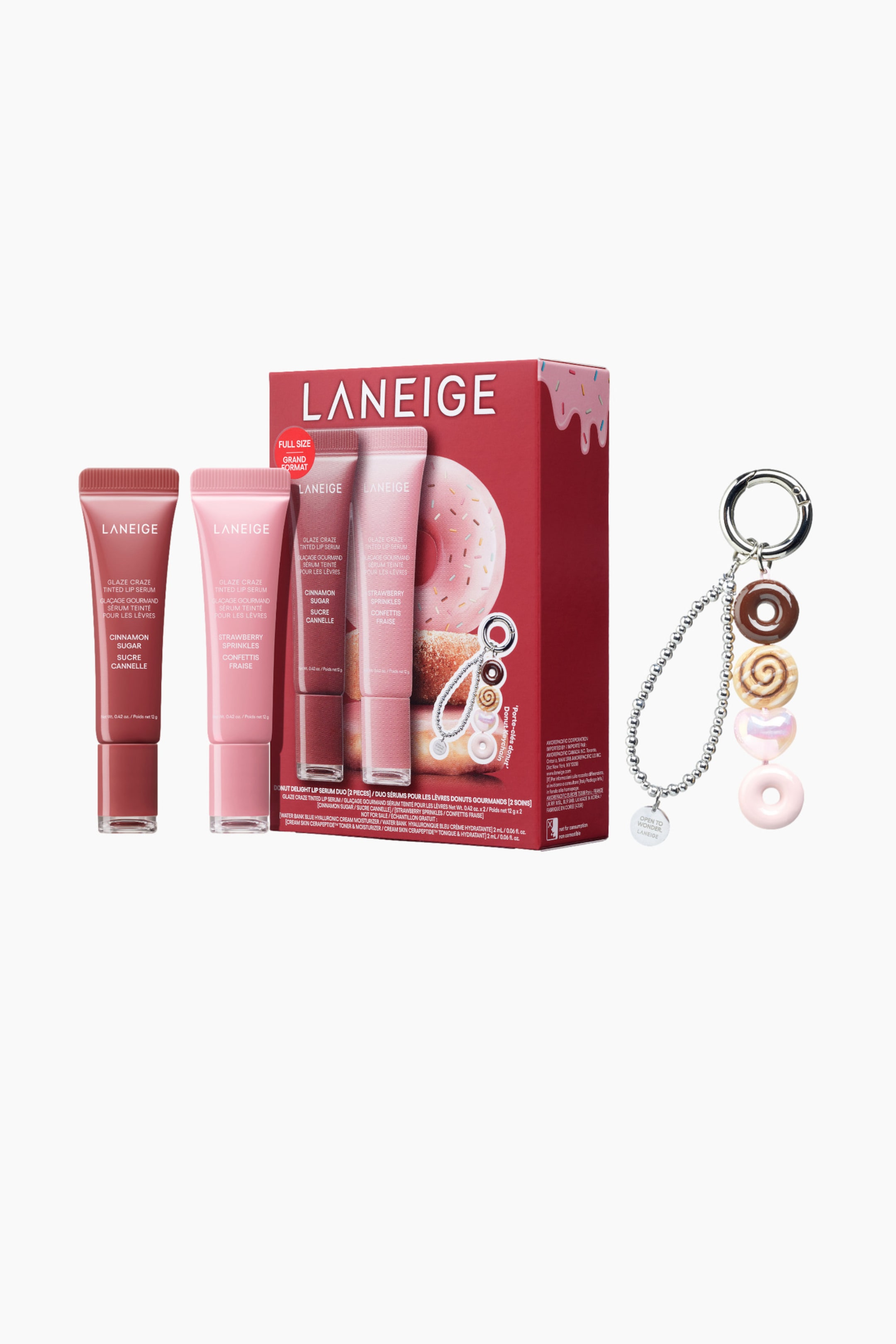 Visa större bild: Ett Laneige Glaze Craze Tinted Lip Serum-set och en nyckelring med donut-tema presenteras. Setet innehåller en röd låda med en rosa donut-grafik, som visar två läppserumtuber i "Cinnamon Sugar" och "Strawberry Sprinkles", med två ytterligare tuber stående upprätt bredvid lådan. Den silverfärgade nyckelringen visar fem donutformade berlocker i nyanser av brunt, iriserande rosa och ljusrosa, hängande från sin ring och pärlkedja.