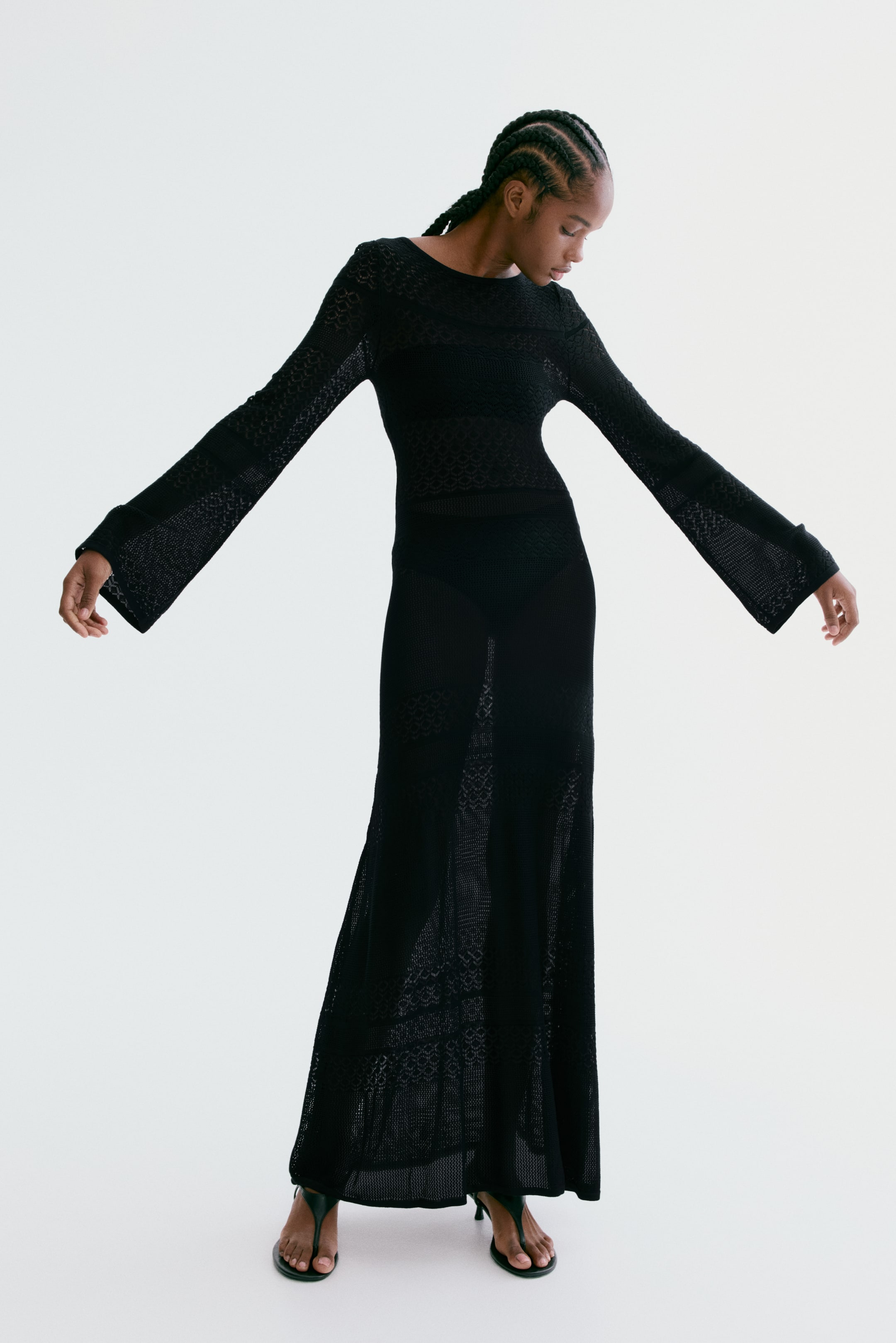 View larger image: A black knit maxi dress featuring a round neckline and long, flared sleeves. The dress showcases intricate openwork patterns of varying density throughout its form-fitting bodice and flowing skirt, creating a textured appearance.