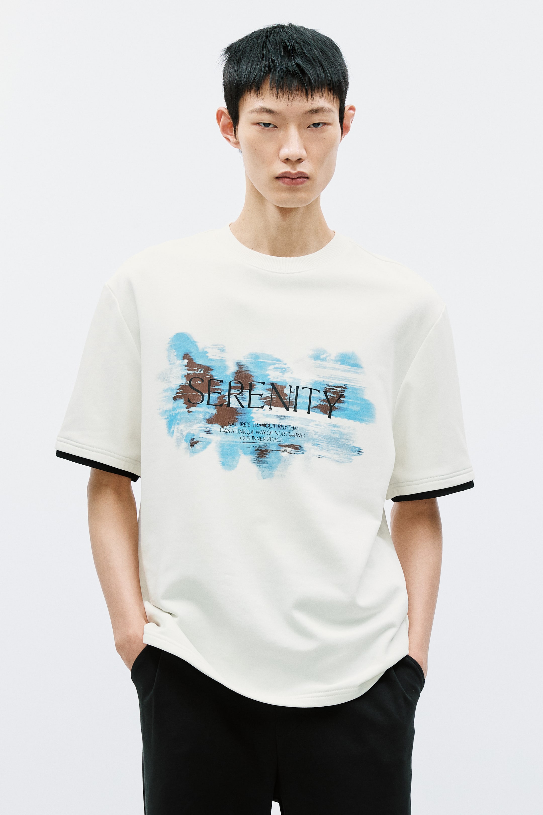 이미지 확대 보기: 오프 화이트 크루 넥 티셔츠가 소개되고 있습니다. 중앙에는 "SERENITY"라는 단어가 어두운 글씨로, 추상적인 파란색과 갈색의 붓터치 위에 배치되어 있으며, 그 아래에 작은 글자가 있습니다. 이 티셔츠는 소매 끝에 검은색 라인이 보이는 짧은 소매 디자인으로 제작되었습니다.