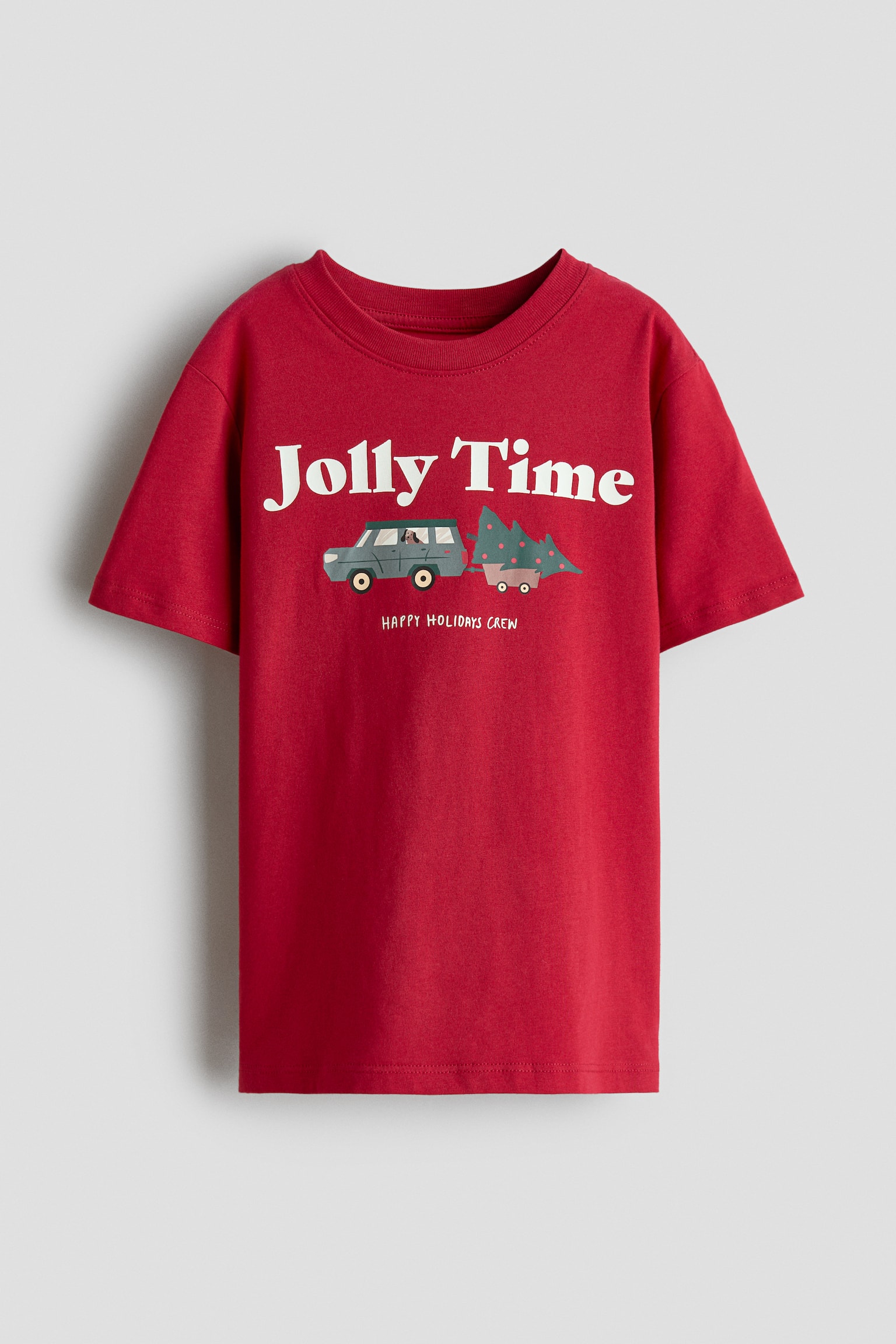 A red crew-neck T-shirt is shown from the front, featuring short sleeves and a straight hem. The chest displays the white text âJolly Timeâ above a graphic of a grey vehicle towing a pine tree in a small trailer, with the words âHAPPY HOLIDAYS CREWâ printed below.