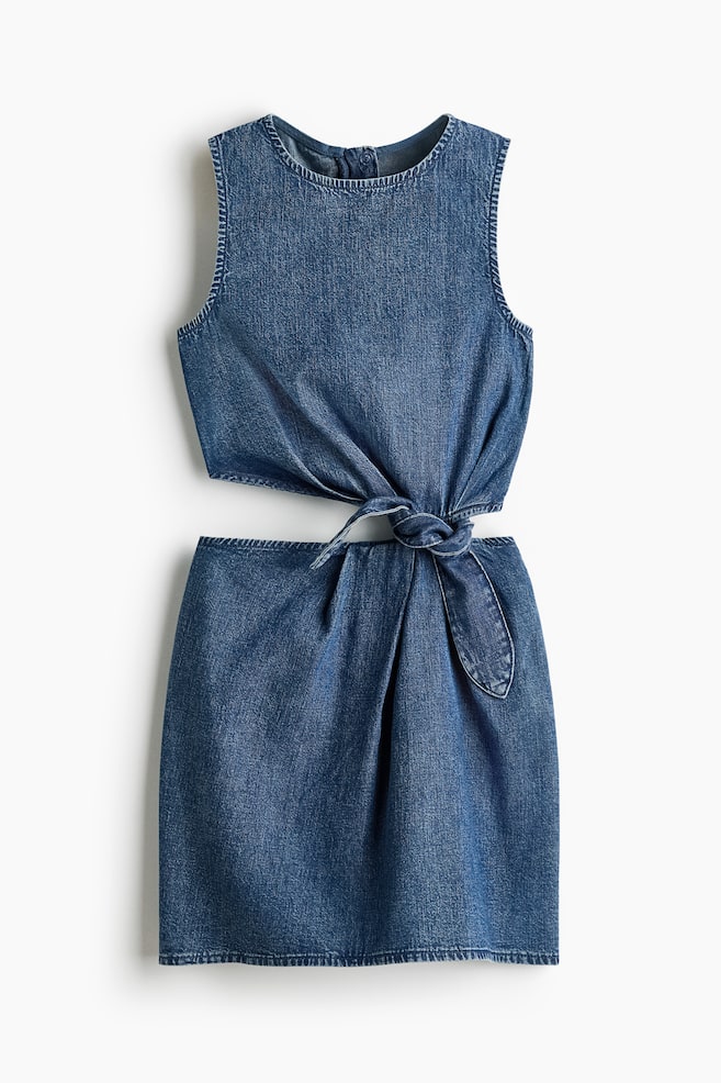 A denim dress is featuring a sleeveless design with a round neckline and a cut-out waist. A tie-knot is connecting the top and the mini-length skirt at the front waist. - 1