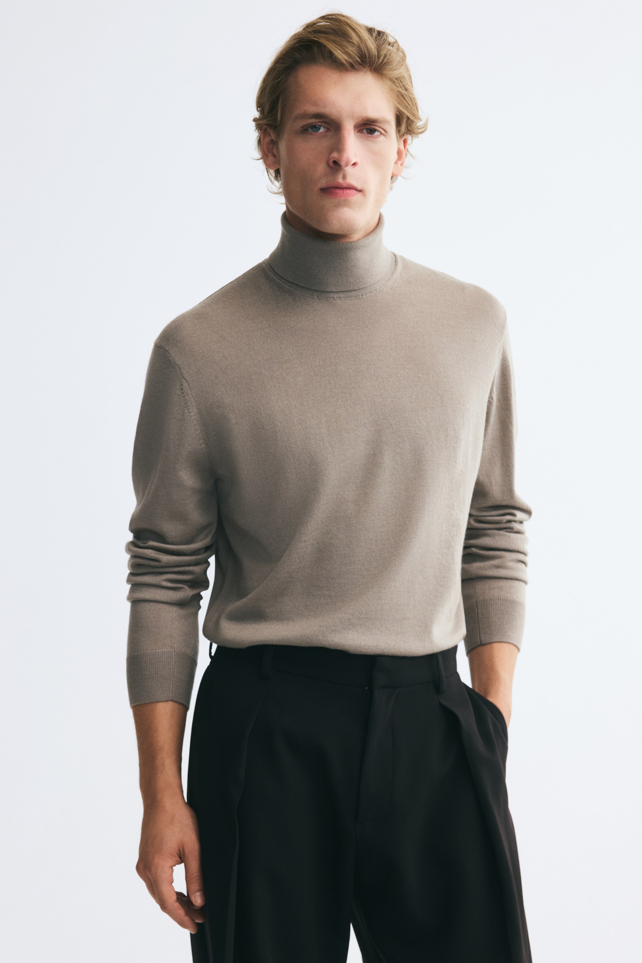 View larger image: A beige fine-knit turtleneck sweater is being worn by the model, styled tucked into black trousers. The sweater features a ribbed turtleneck collar, gently dropped shoulders, and long sleeves with ribbed cuffs.