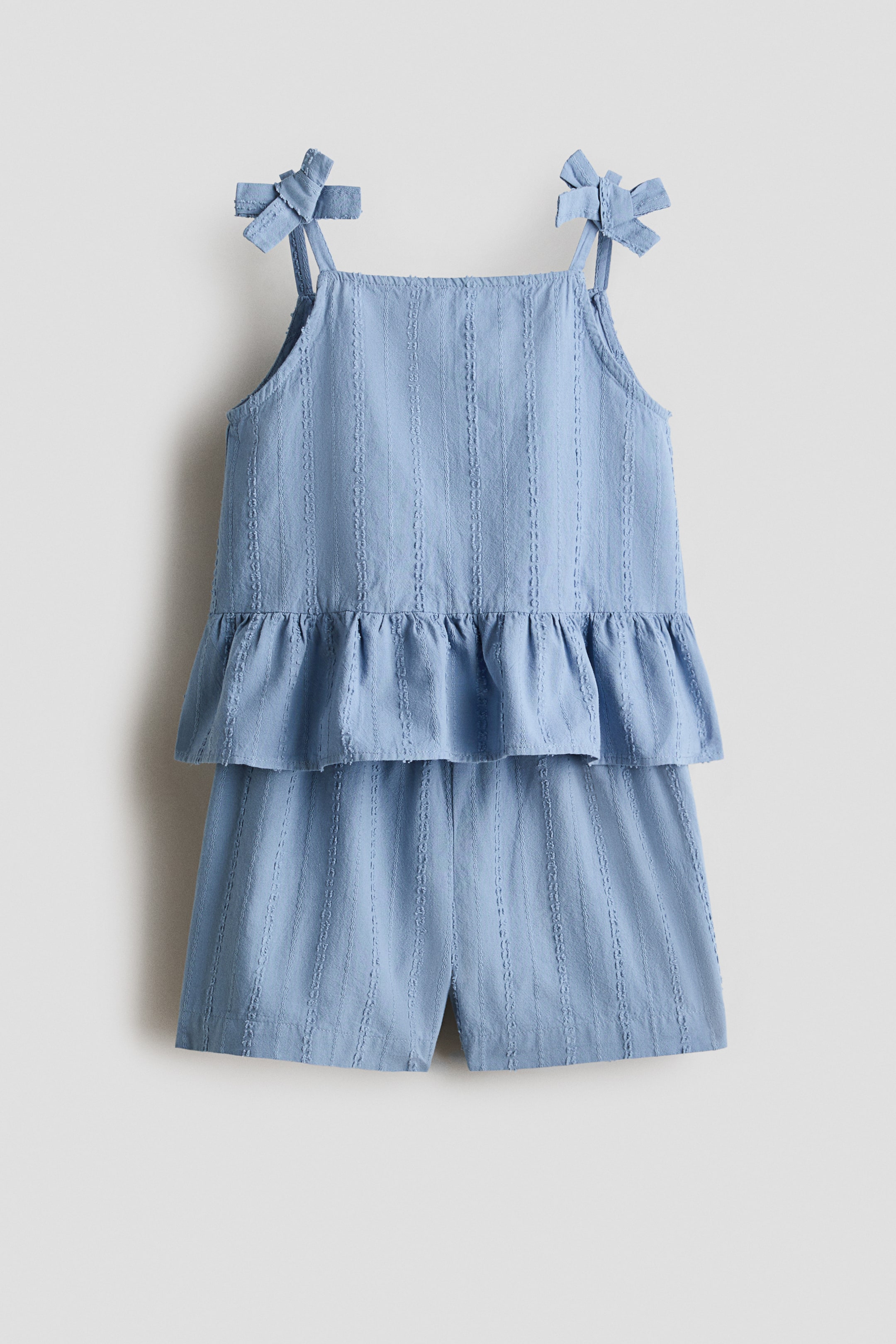 View larger image: A light blue two-piece set is shown from the front, featuring a top and matching shorts. The top has a square neckline, thin spaghetti straps tied into bows at the shoulders, and a peplum hem. Both pieces display a pattern of vertical textured stripes.