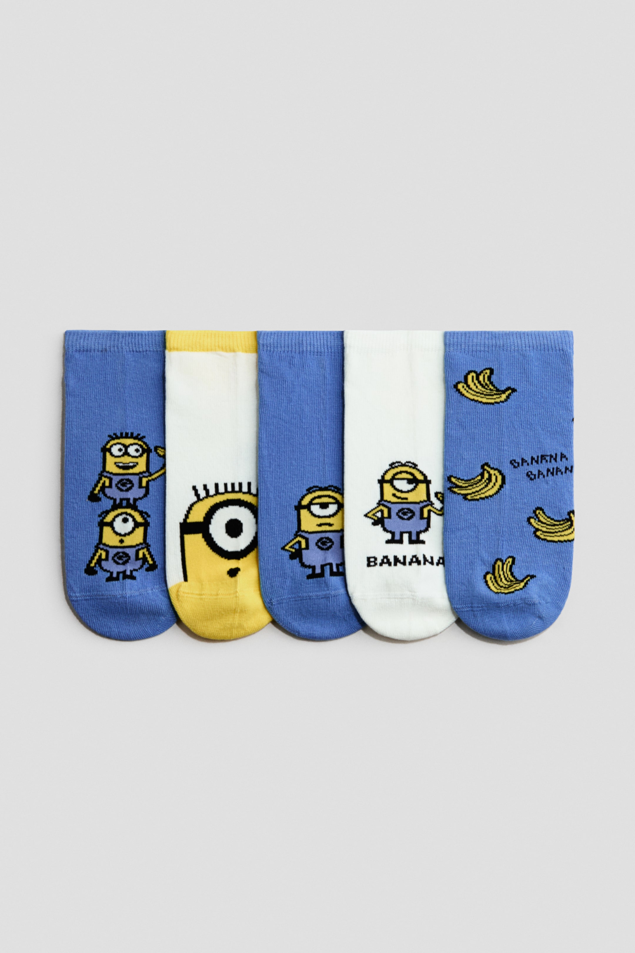 Agrandir l'image: Cinq chaussettes de sport en maille jacquard dans un mélange doux de coton avec élastication autour du haut sont posées à plat, affichant leur devant. Elles présentent des motifs bleus et blancs avec des personnages Minions jaunes et des motifs de bananes.